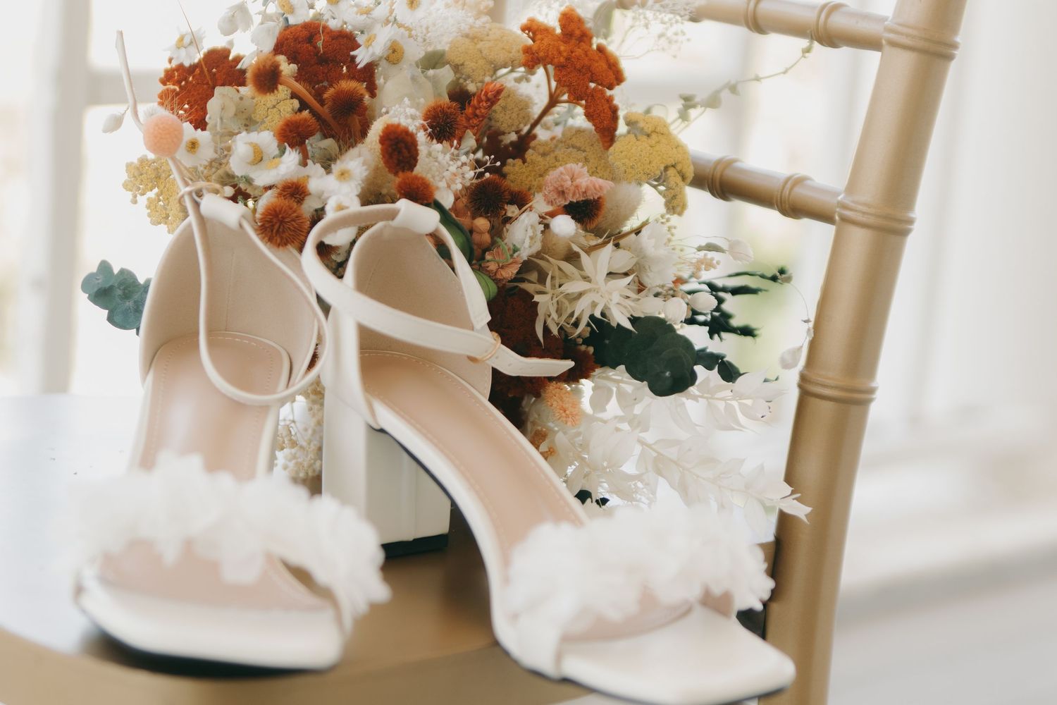 White bridal shoes and floral arrangements displayed on wooden chairs.