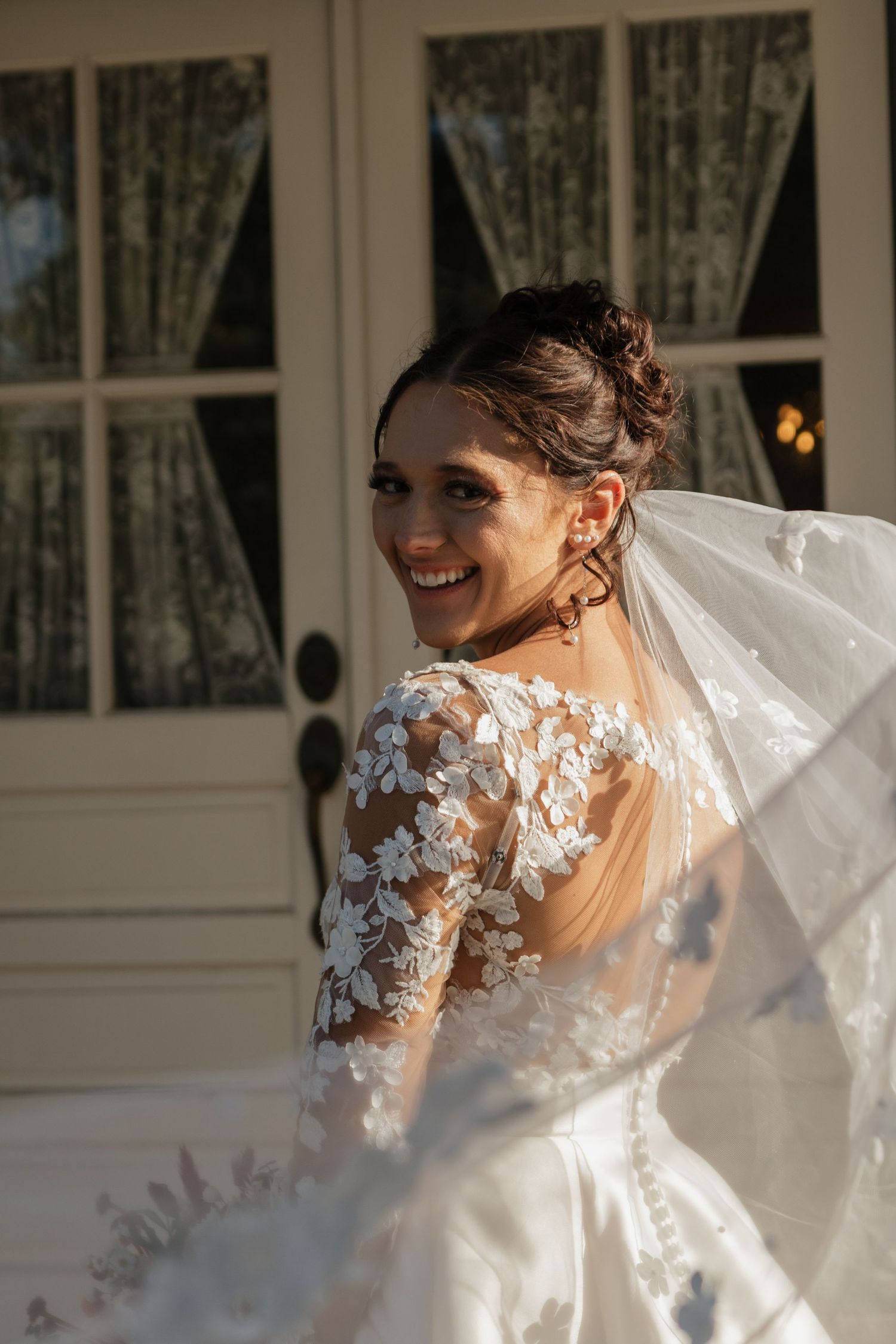 A bride in a lace wedding gown twirls her dress in front of a window.
