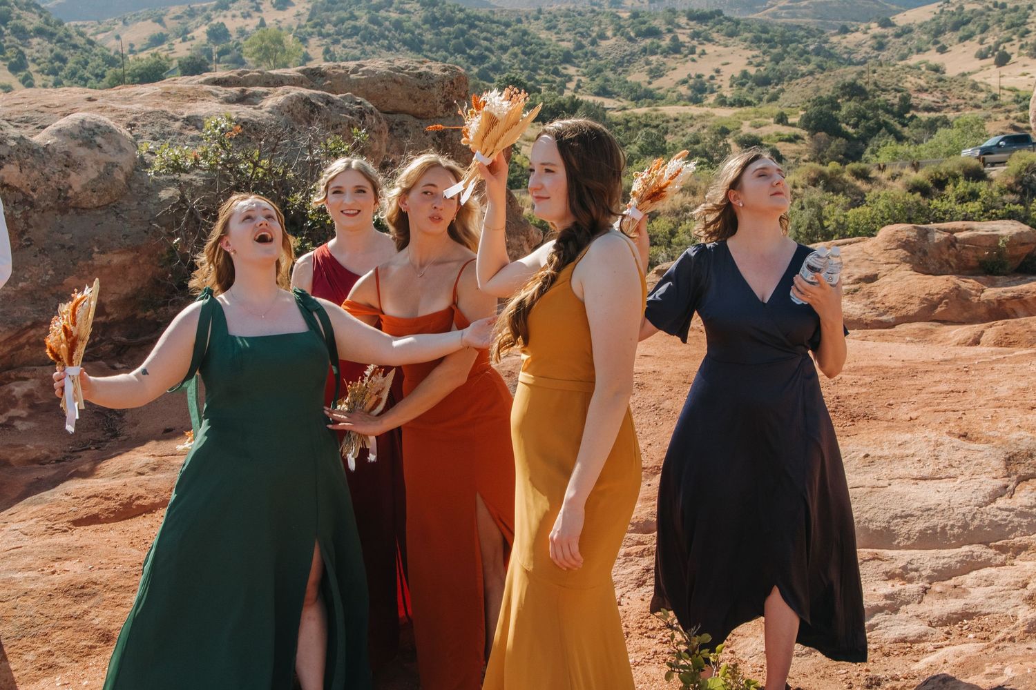 Bridesmaids in colorful dresses celebrate joyfully on red rocks in a scenic mountain setting.