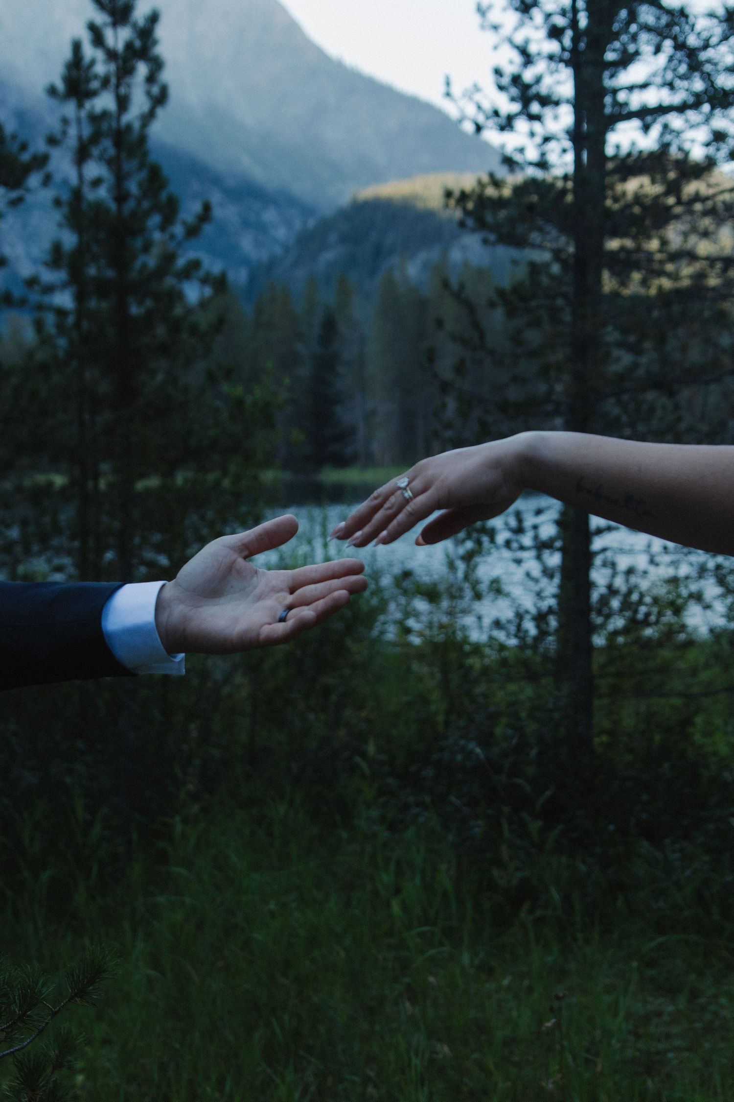Two hands reach toward each other against mountain lake sunset backdrop.