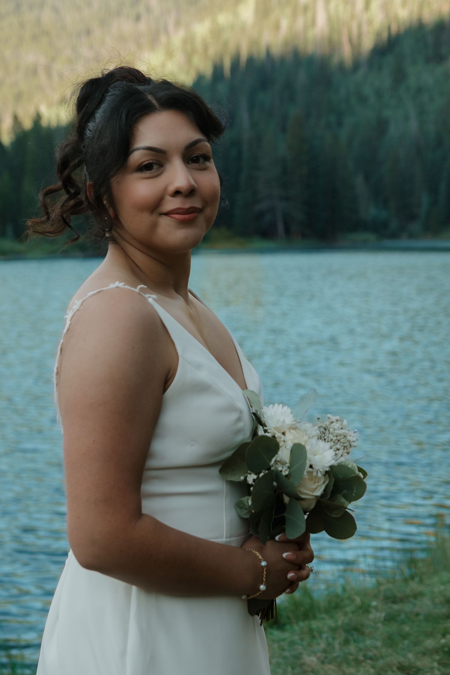 Bride in elegant white dress holds a classic white and green bouquet by the lake.
