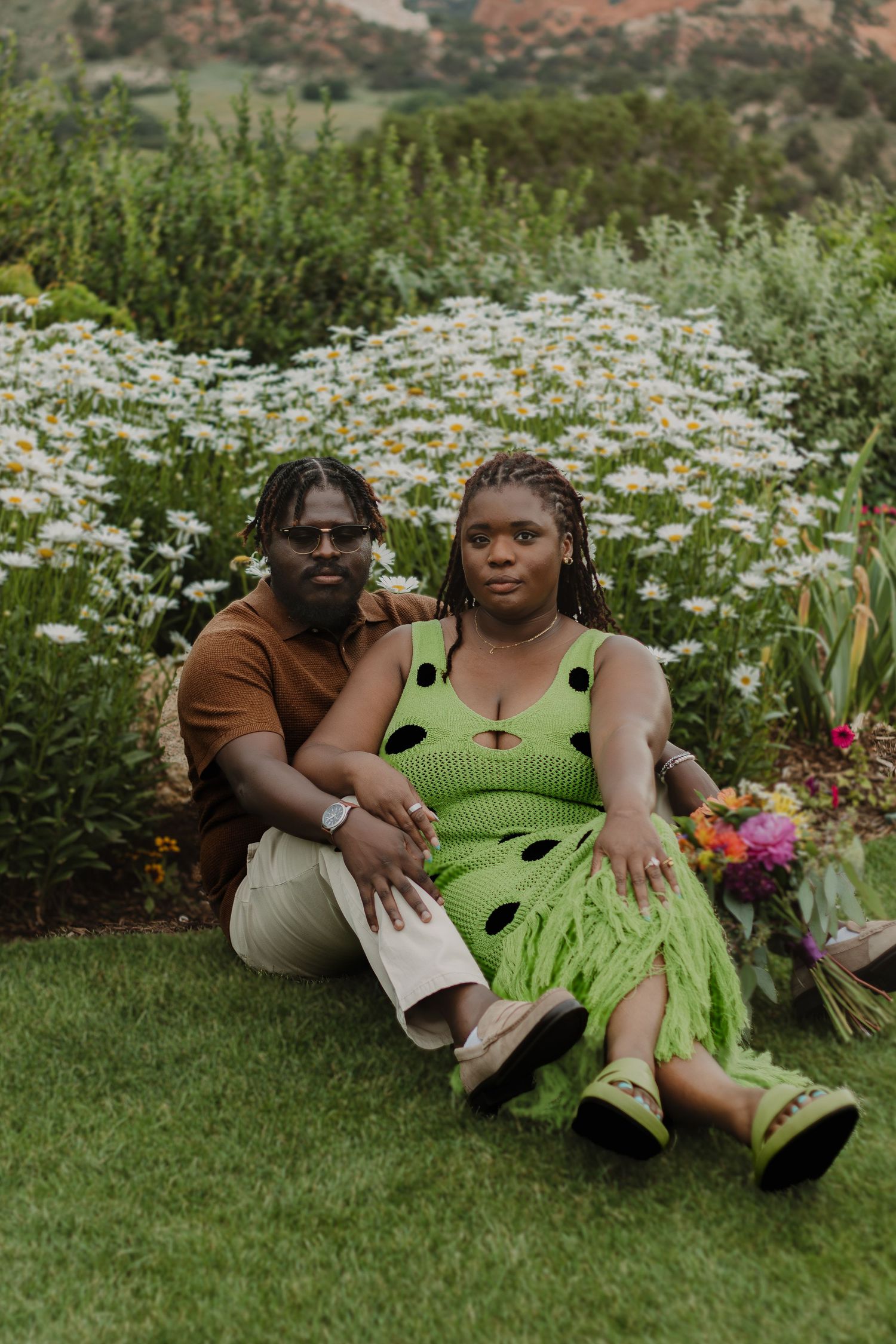 Couple sitting together on grass surrounded by blooming daisies and greenery in a garden setting.