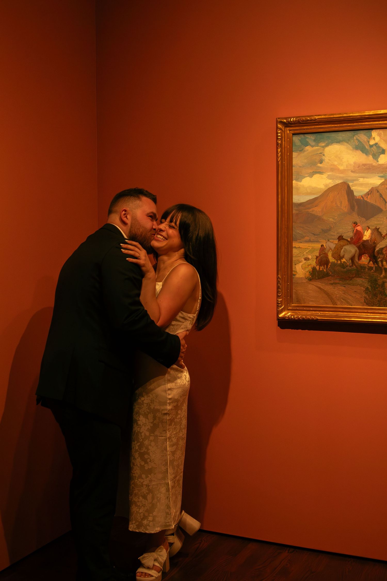 Couple embracing in moody lighting against red gallery wall with framed mountain artwork.