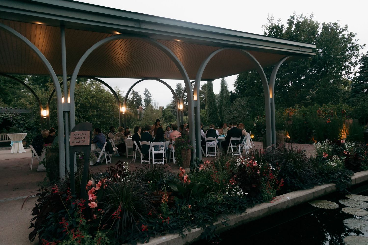 Romantic outdoor wedding reception under a lit garden pavilion surrounded by colorful flowers at dusk.