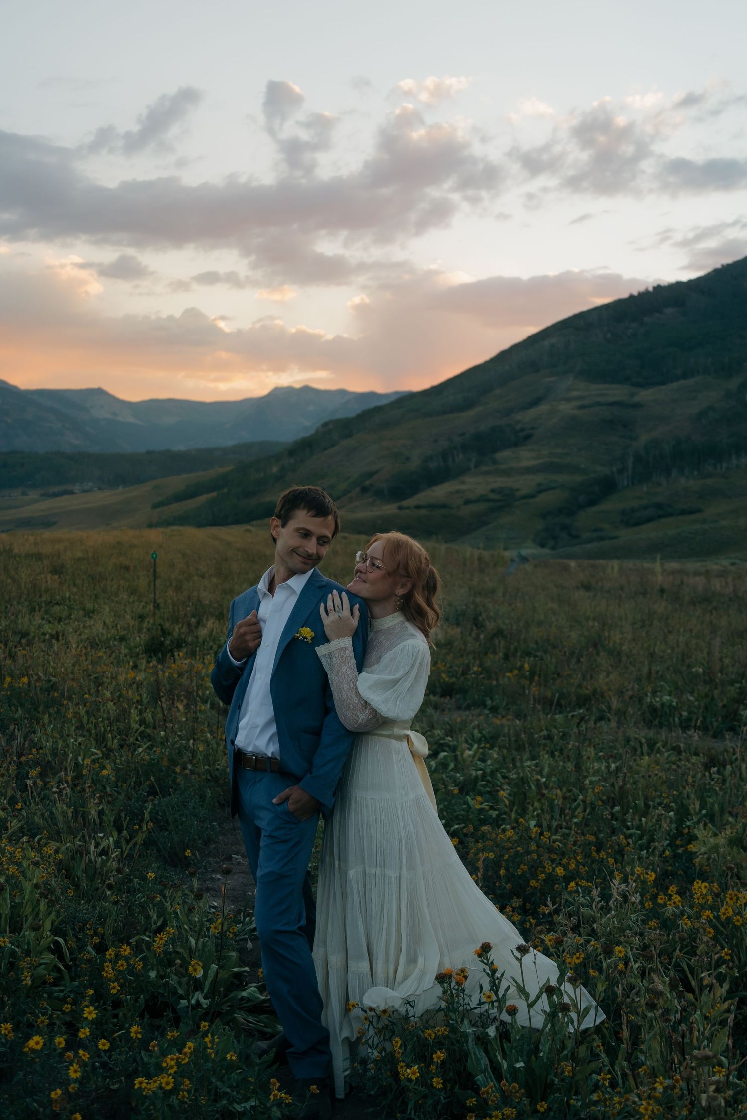 Bride and groom embracing in wildflower field at sunset with rolling mountain hills behind them.