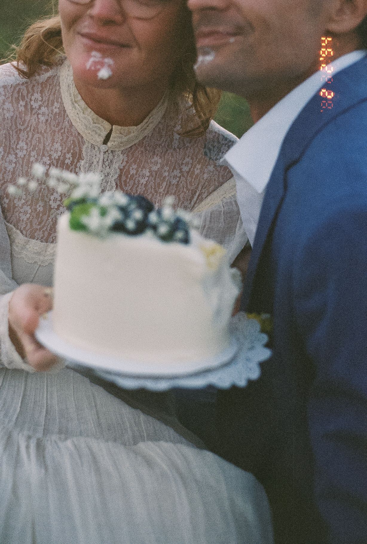 Bride and groom holding white wedding cake decorated with blueberries and flowers outdoors.