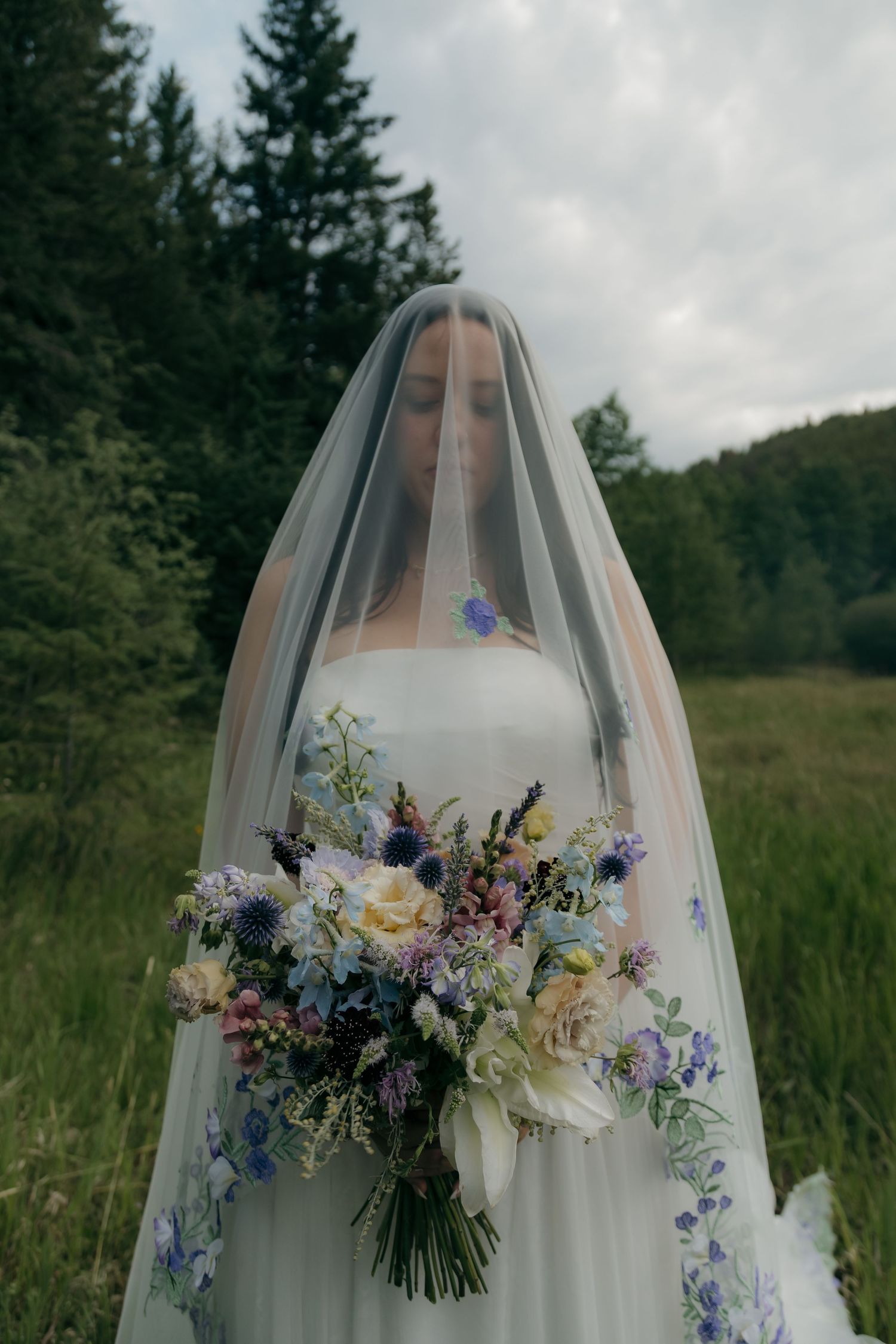 Bride veiled in sheer fabric holds colorful wildflower bouquet with blue and purple blooms outdoors.