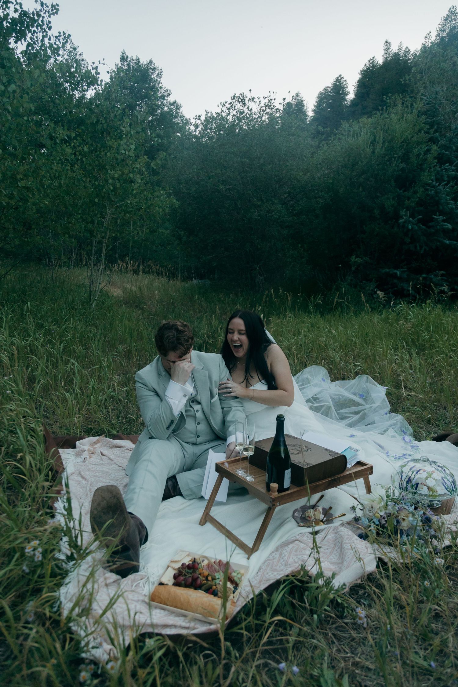 Newlywed couple laughing during romantic outdoor picnic on white blanket in a forest clearing.