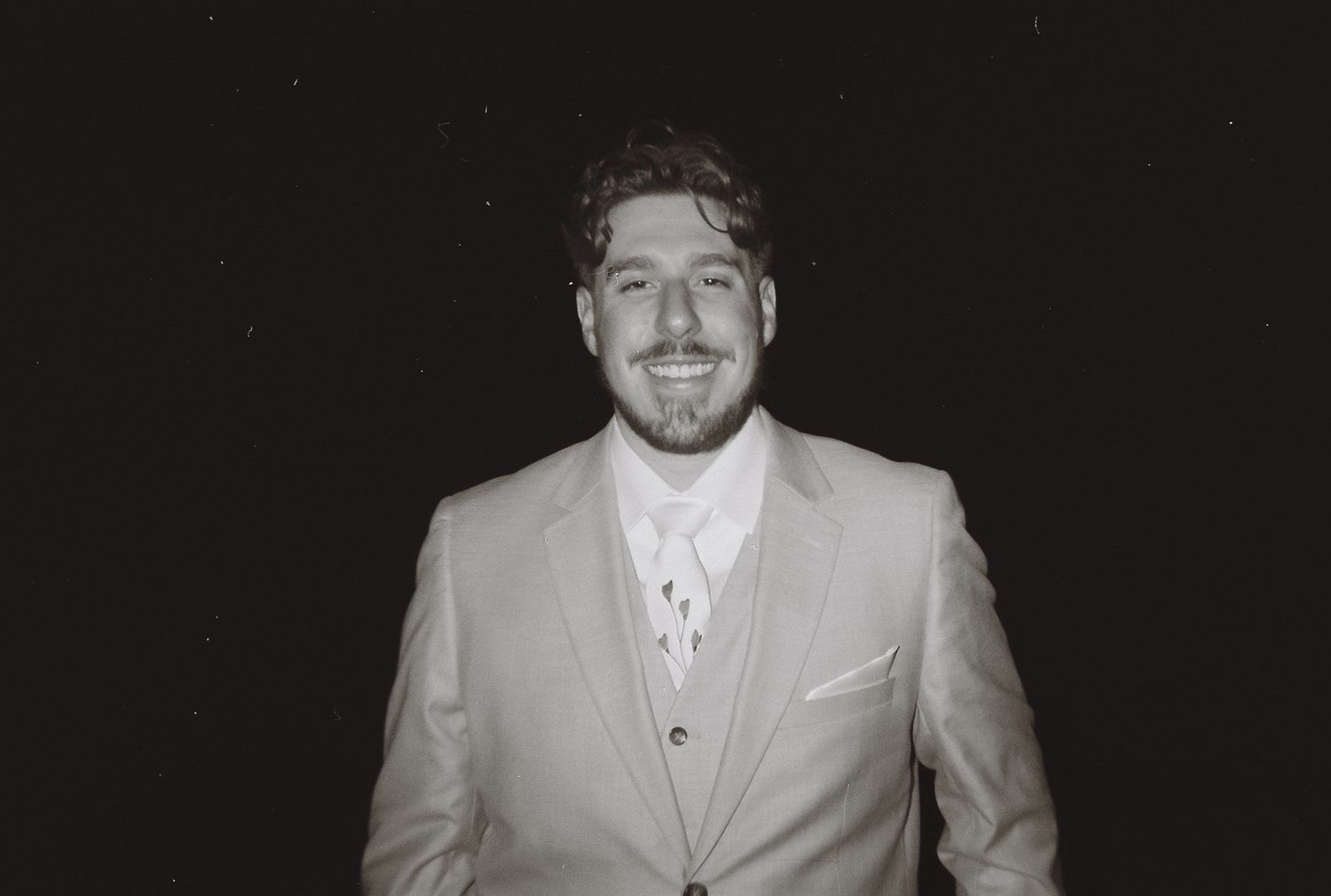 Black and white portrait of smiling groom in light gray suit at night.