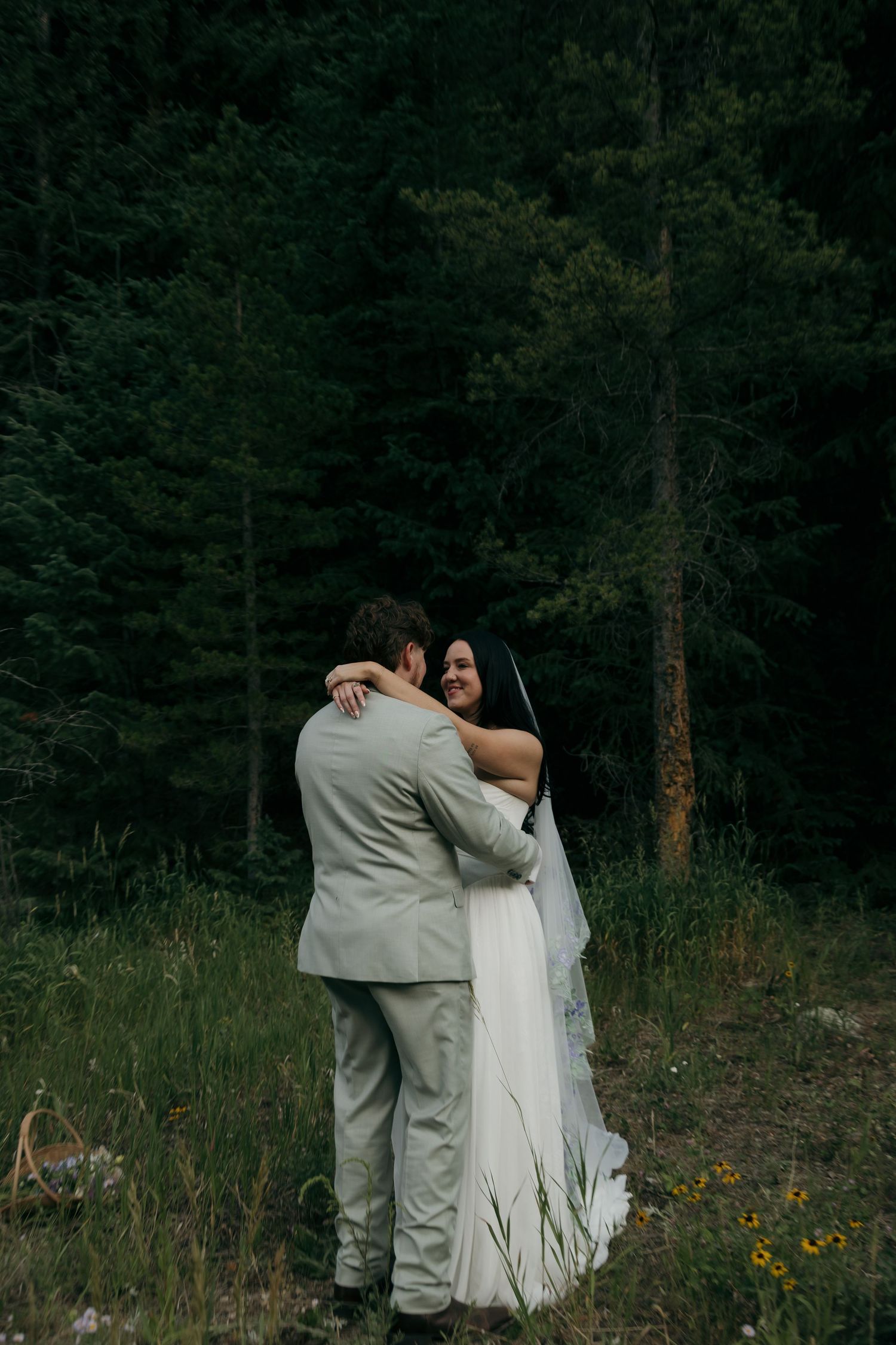 Smiling bride hugs groom in forest meadow with wildflowers during outdoor mountain elopement.