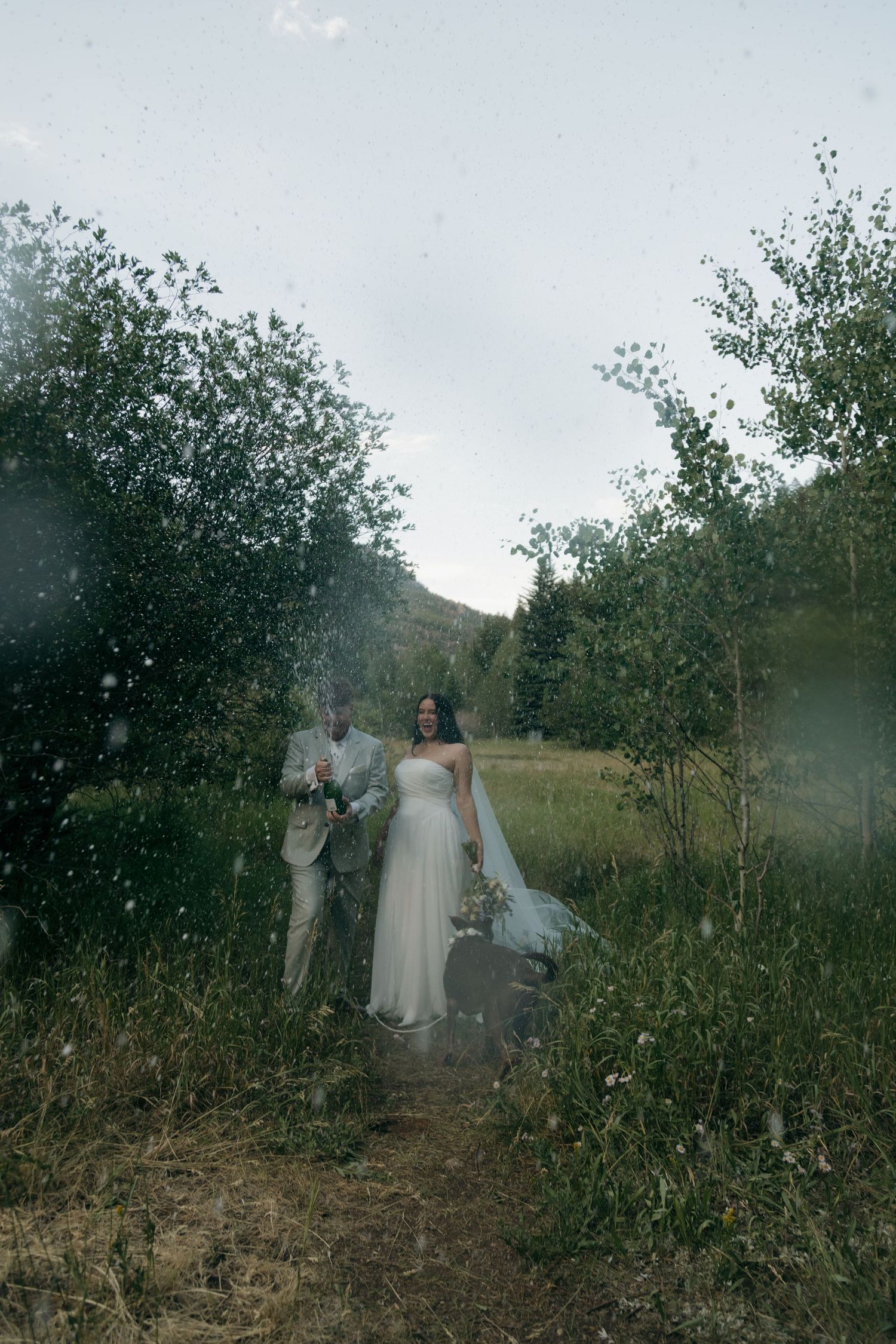 Bride and groom celebrating with champagne spray on a scenic outdoor trail surrounded by lush green trees and mountains.