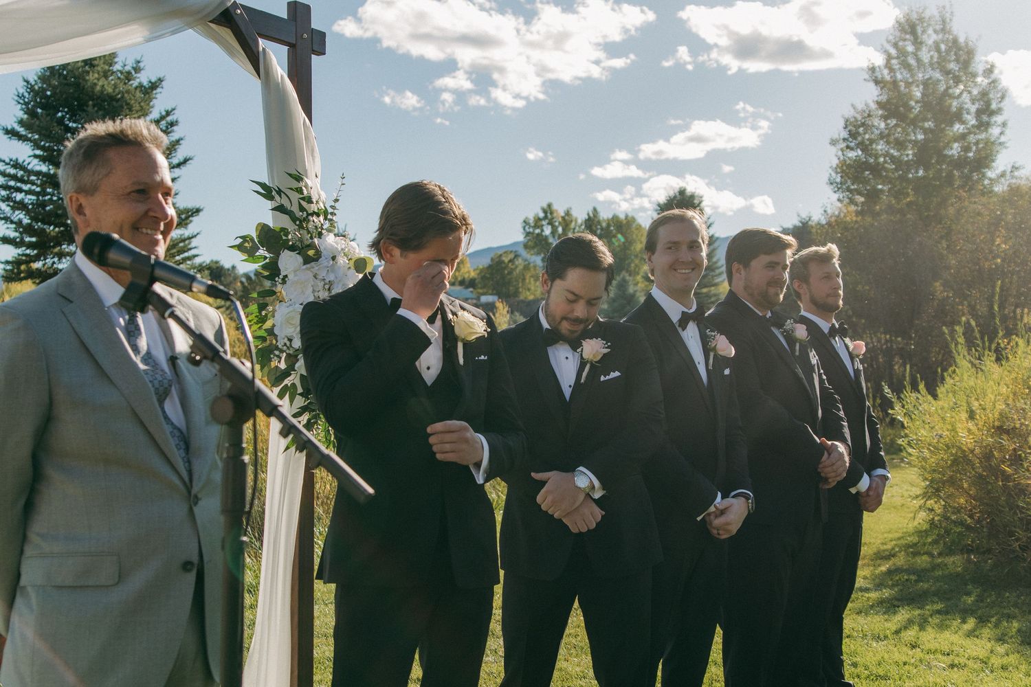 A group in formal attire stands outdoors at a wedding ceremony during golden hour lighting.