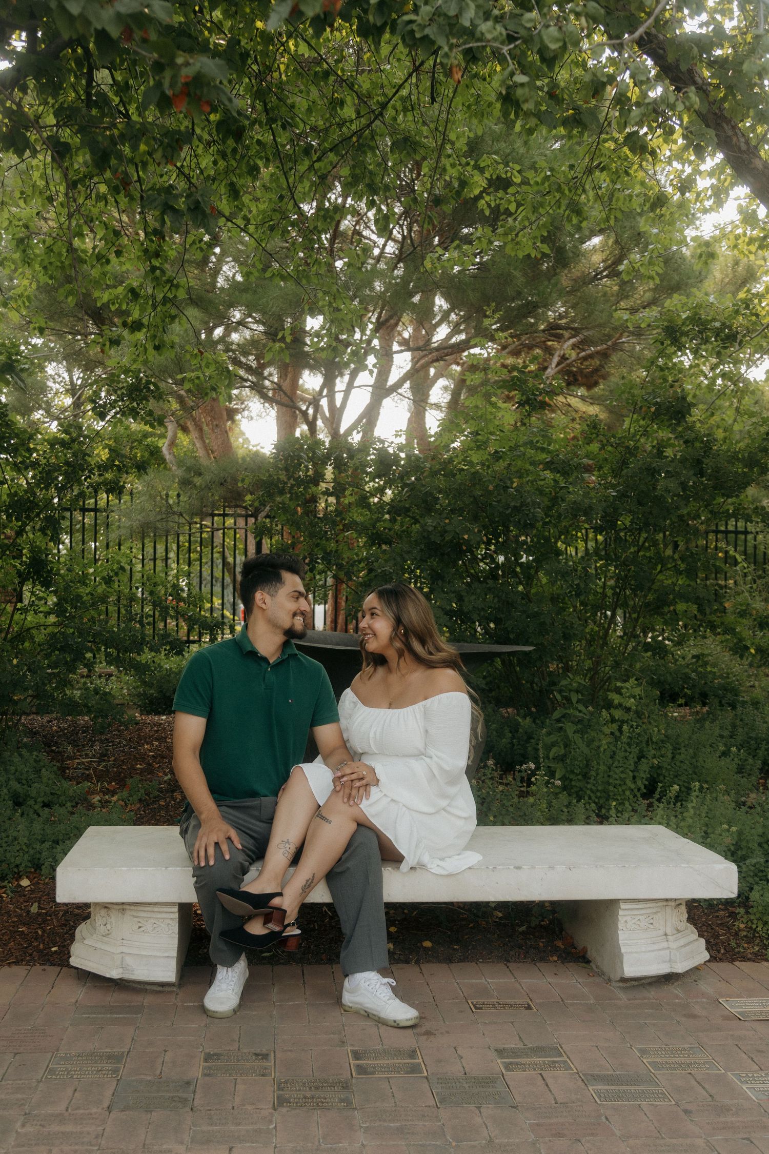 Couple sitting together on a white stone bench in a lush green garden, gazing at each other.