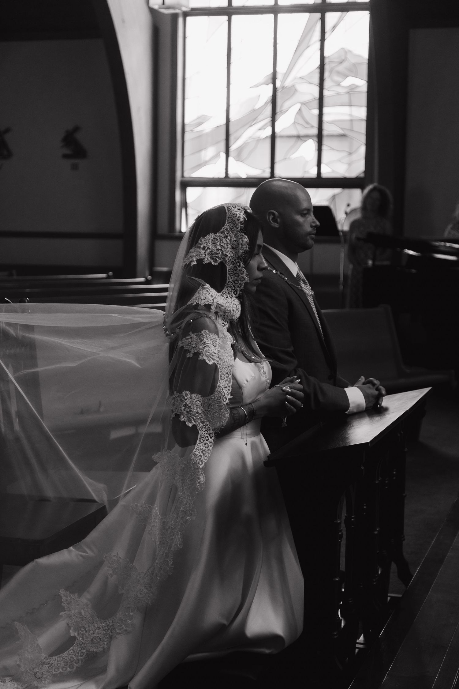Black and white photo of bride and groom kneeling in prayer at church pew during ceremony.