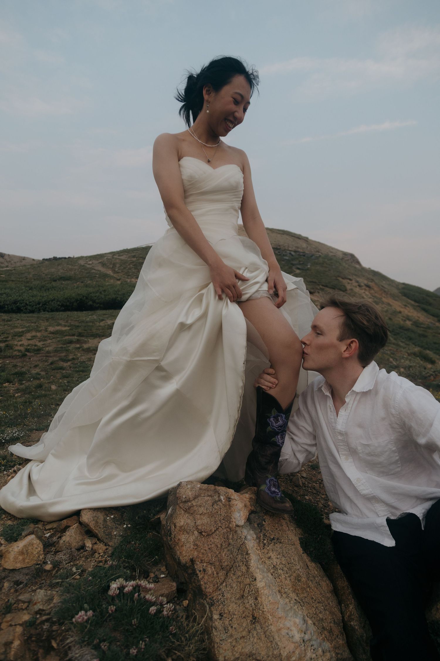 Groom in white shirt kisses bride's leg as she sits elevated on a rock in a mountain landscape.