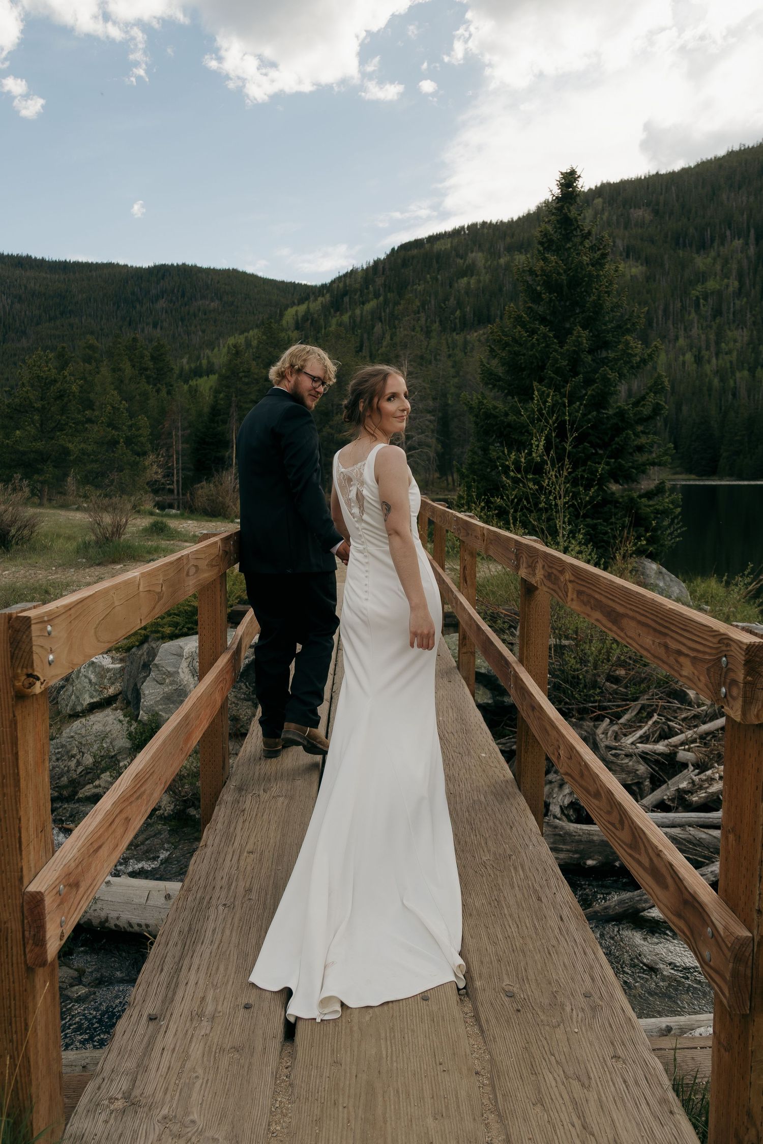 Bride and groom walk wooden bridge hand in hand with scenic mountain lake backdrop.