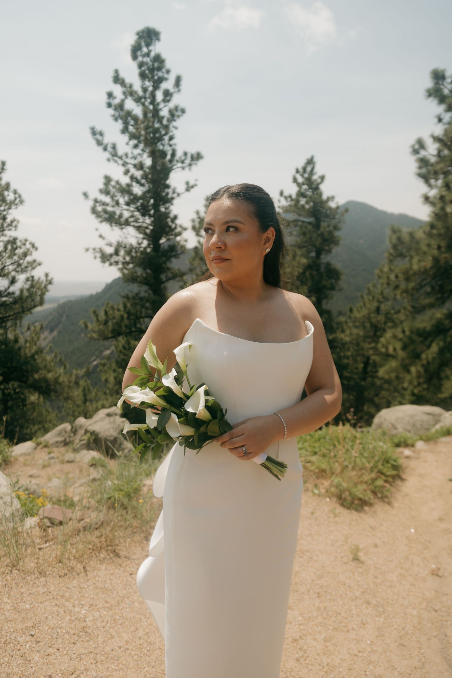 Bride in white strapless wedding gown holding calla lily bouquet on a mountain trail.