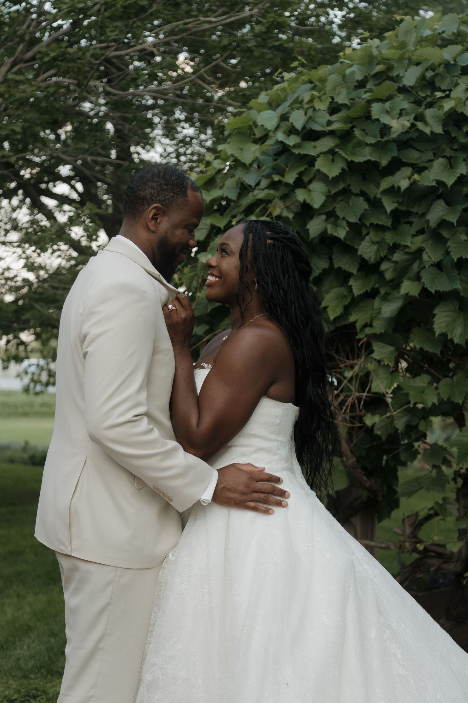 Smiling Black bride in a strapless white gown embracing her groom in a cream suit before lush green vines.