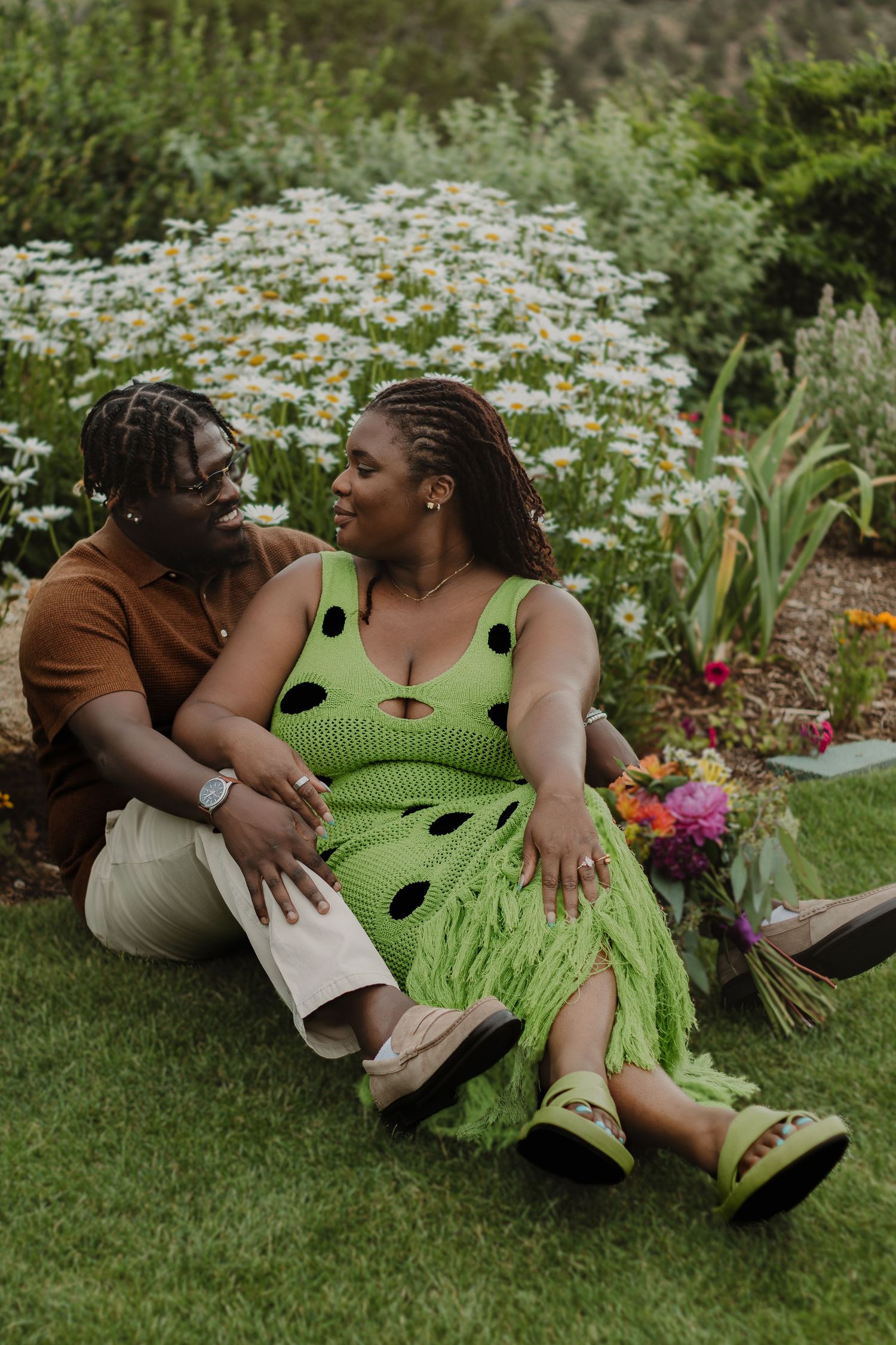 A couple shares an intimate moment while sitting on grass surrounded by blooming daisies in a garden setting.