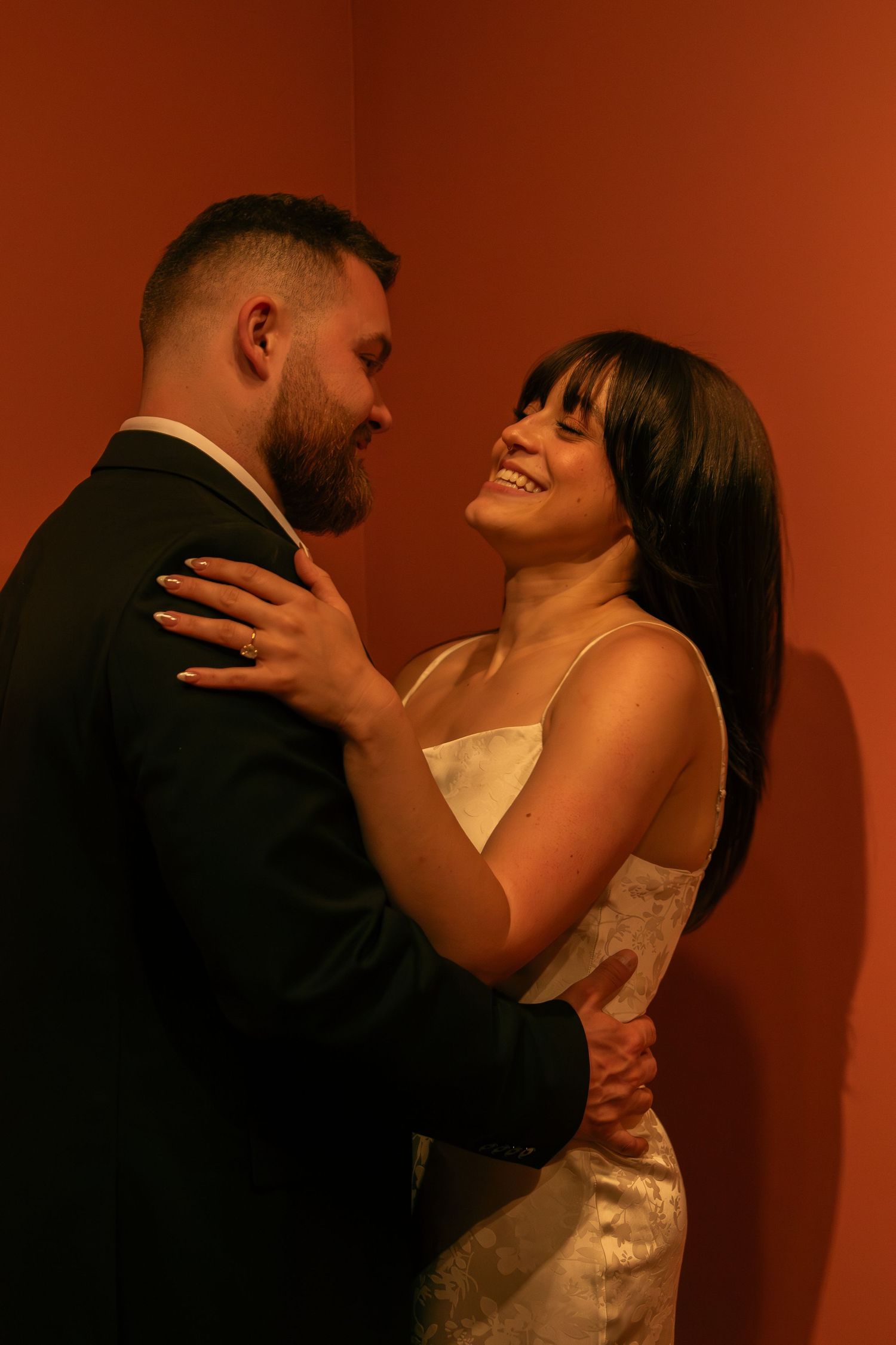 A couple embraces in formal attire against a coral-colored wall in warm lighting.