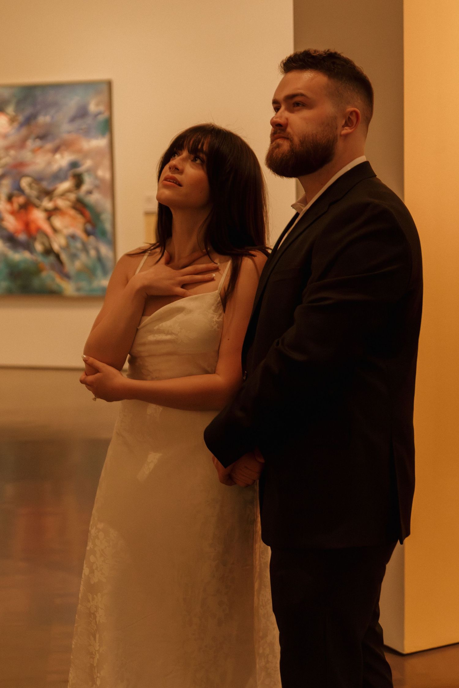 Two figures embrace in an art gallery with paintings visible on the walls in warm lighting.