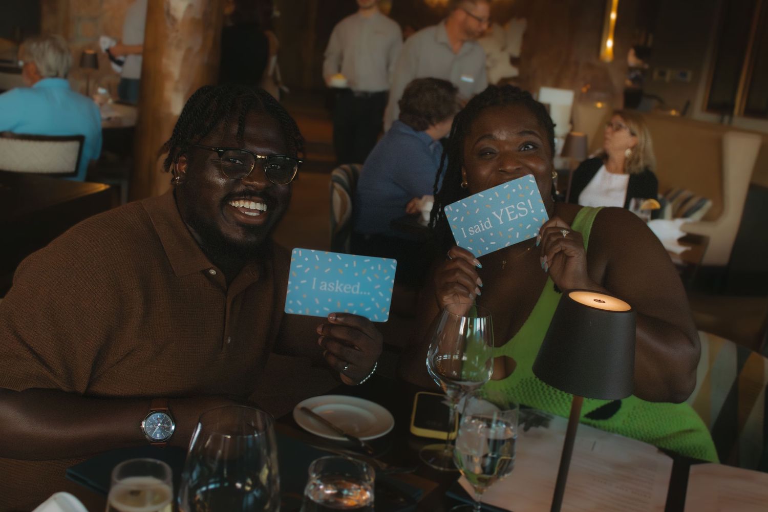 Two friends smile while holding up blue tickets during a dinner gathering at a restaurant.