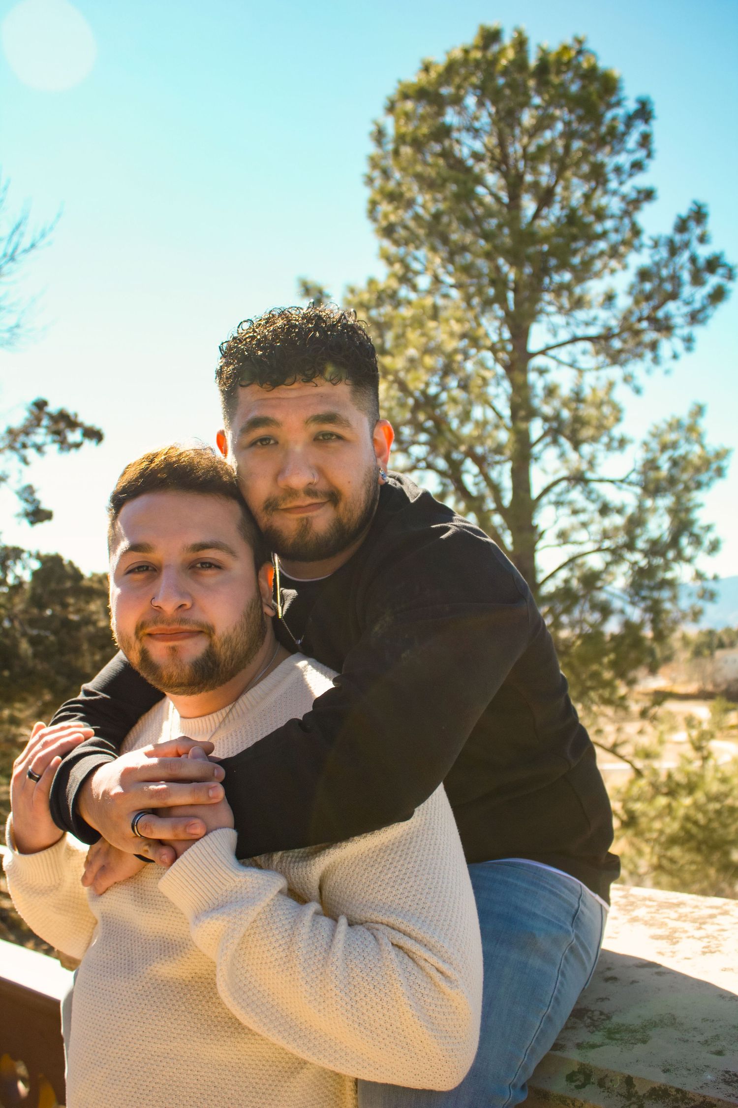 Two people embrace outdoors with pine trees and blue sky in the background.