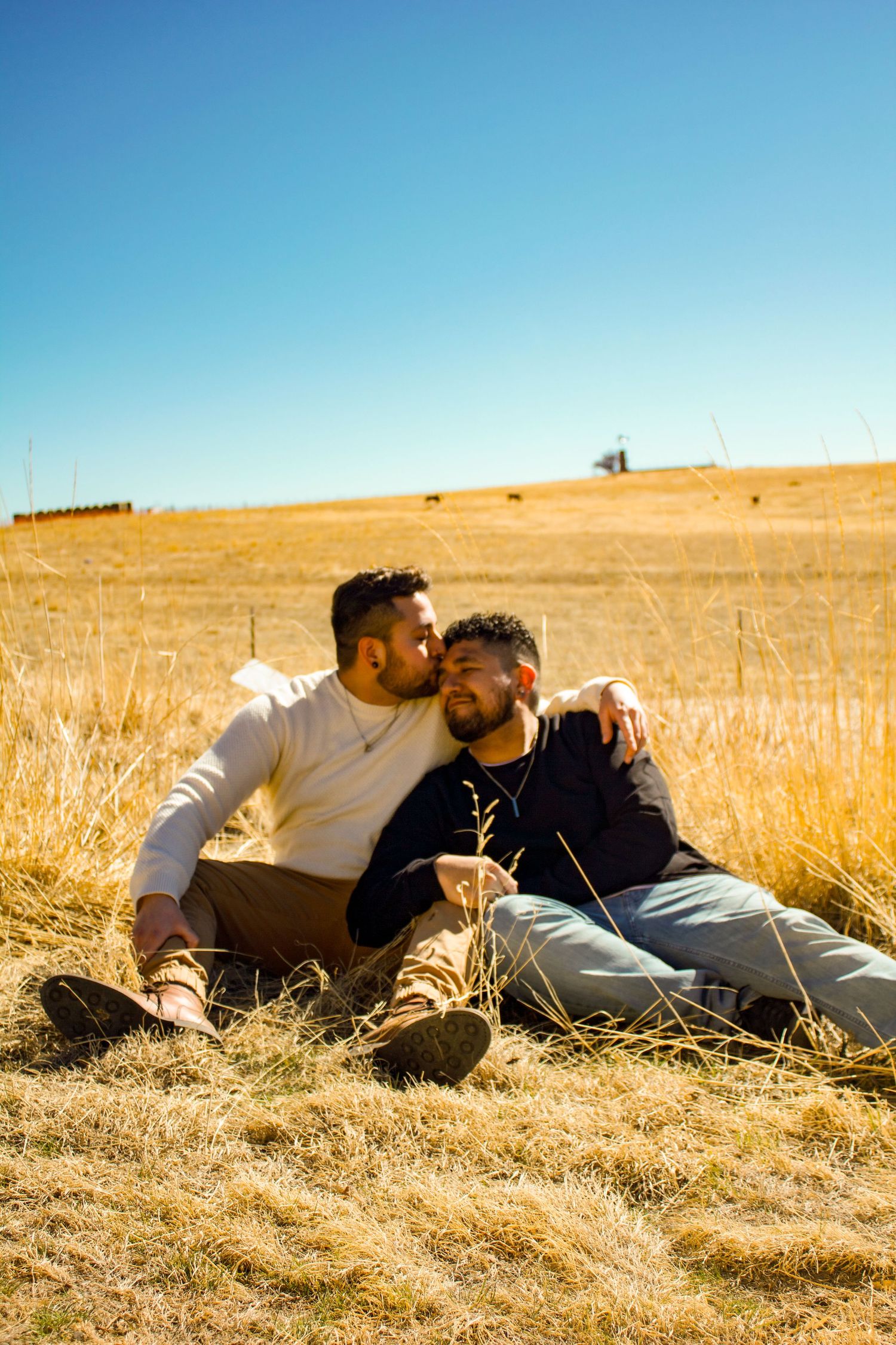 Couple embracing while sitting in dry grass field with blue sky backdrop.