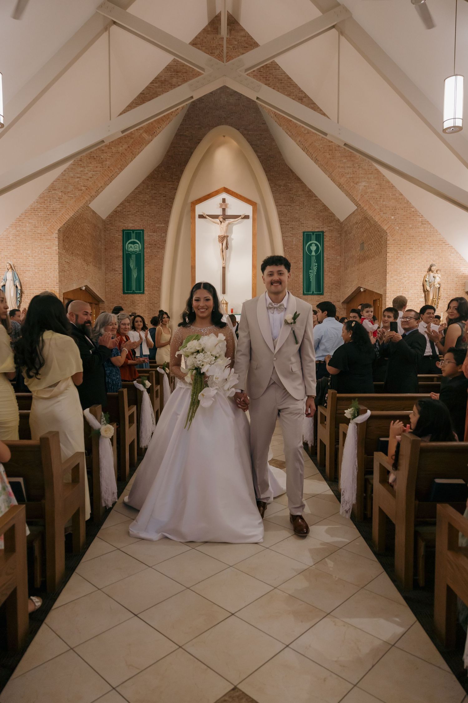Newlyweds walking joyfully down church aisle after Catholic wedding ceremony with guests applauding.