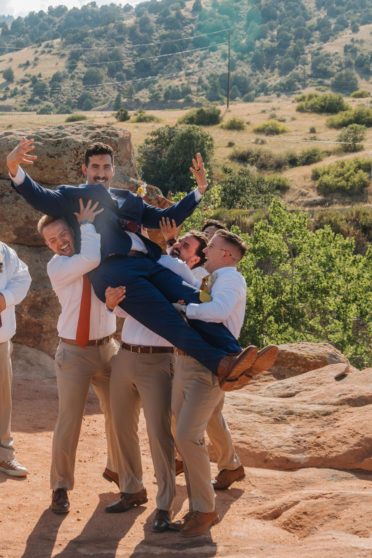 Groomsmen in khaki pants playfully pose on red rocks during a wedding photoshoot.