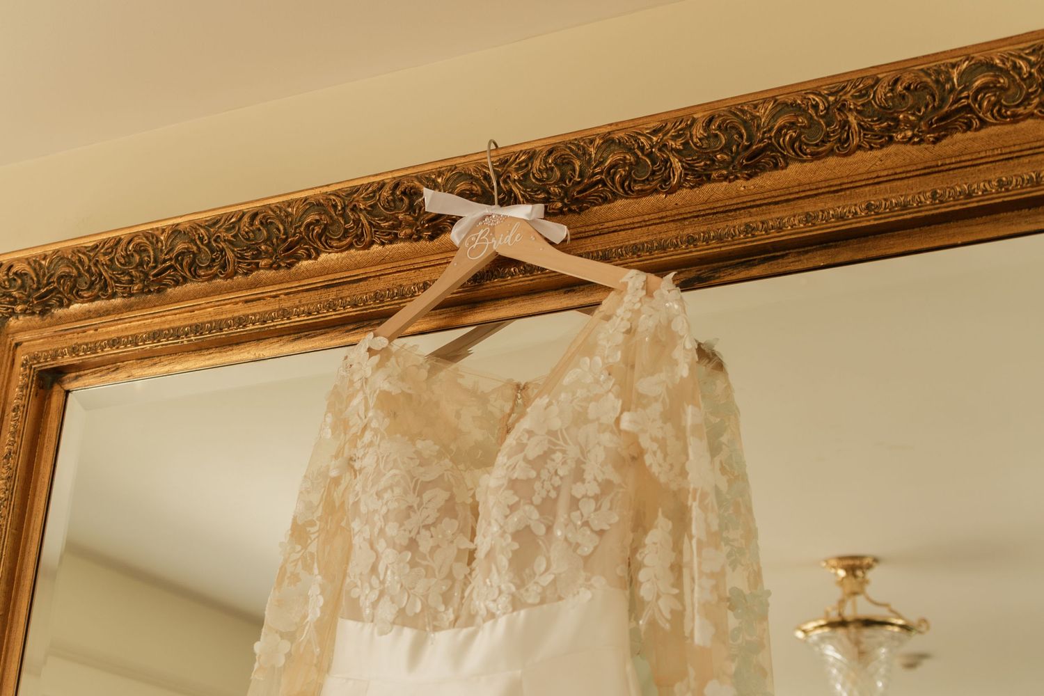 Ornate wedding dress hanging beneath decorative gold-framed molding on a cream-colored wall.