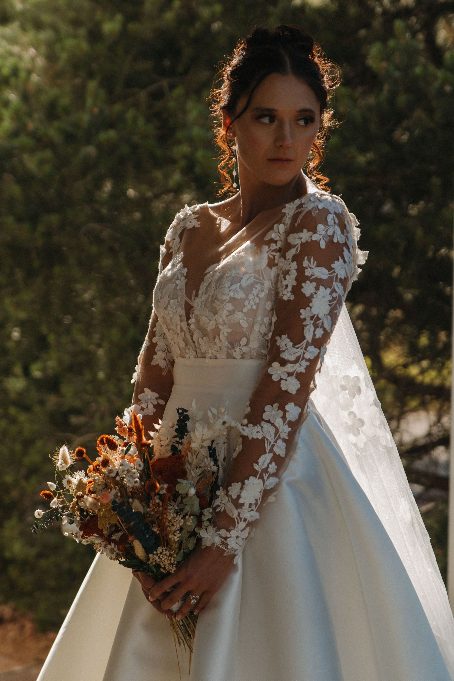 Bride in lace-detailed wedding gown holding autumn bouquet in golden sunlight.