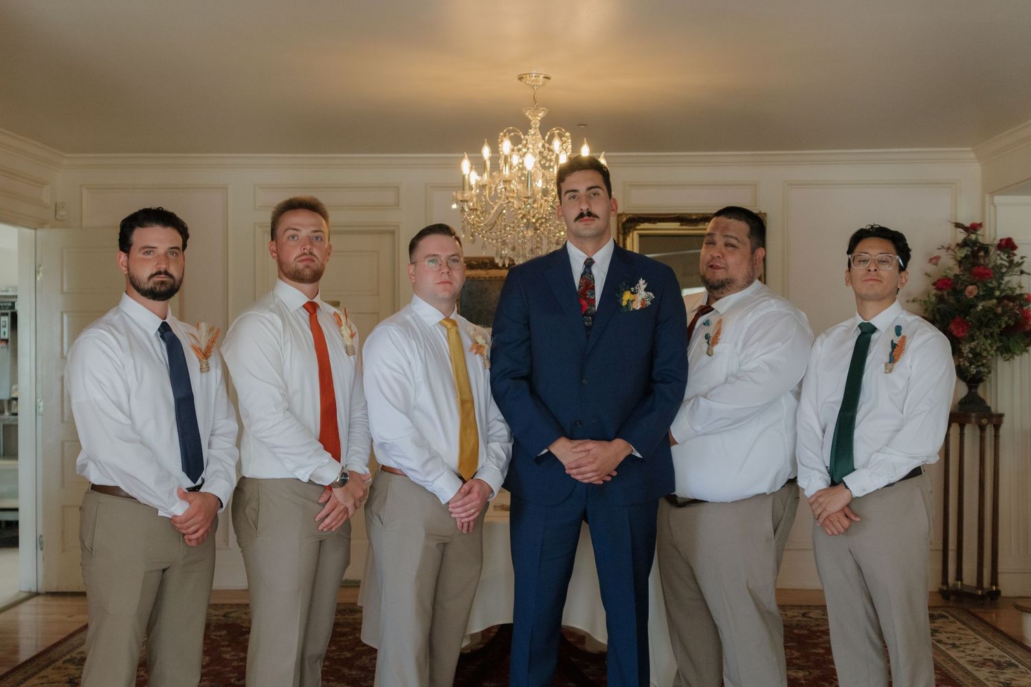 Groomsmen in light suits and ties posing beneath an elegant chandelier.