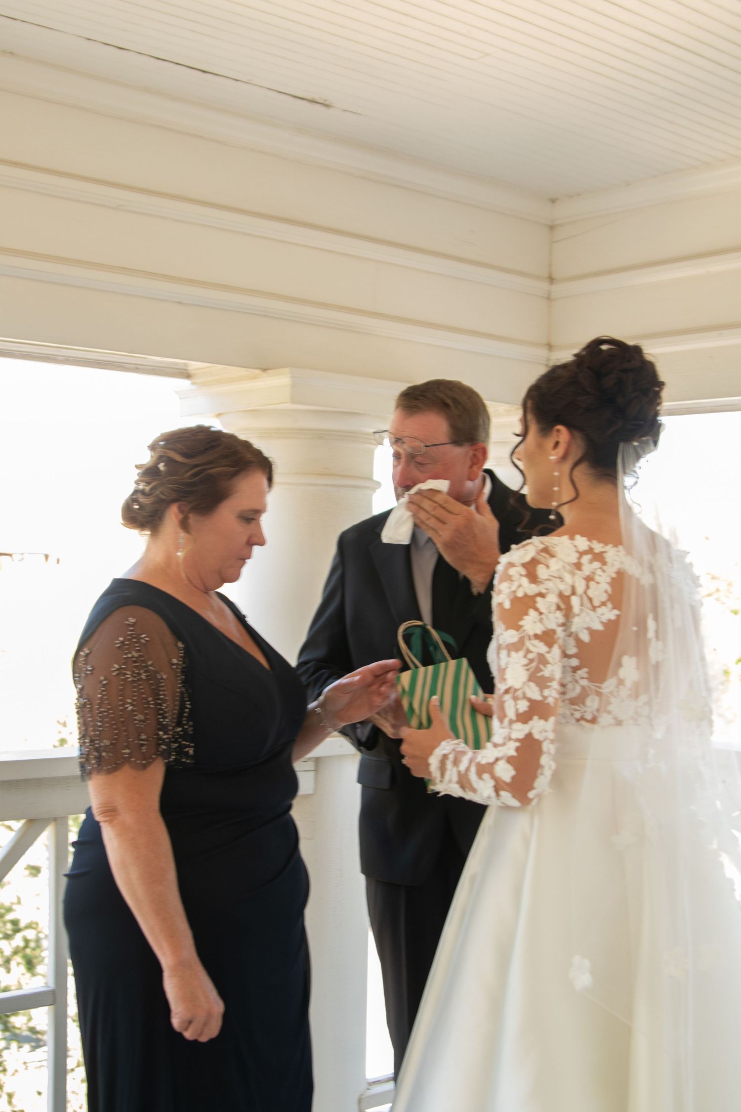 Group of attendants in dark dresses examining and helping with a white wedding gown on a porch.