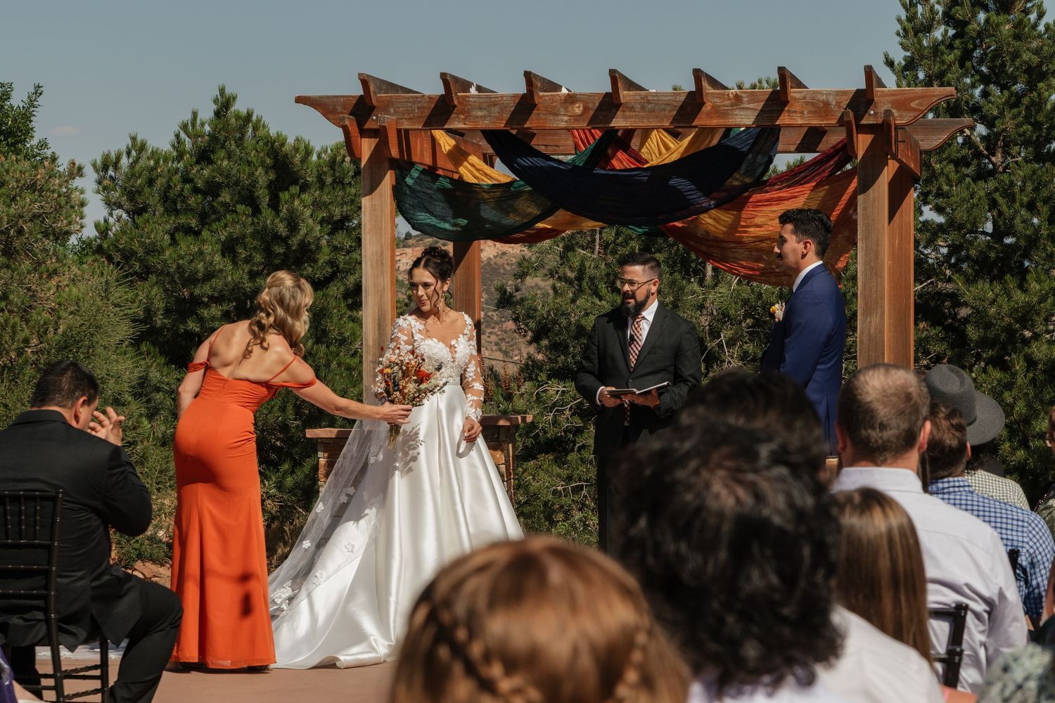 Wedding ceremony taking place under wooden pergola with gathered guests seated outdoors.