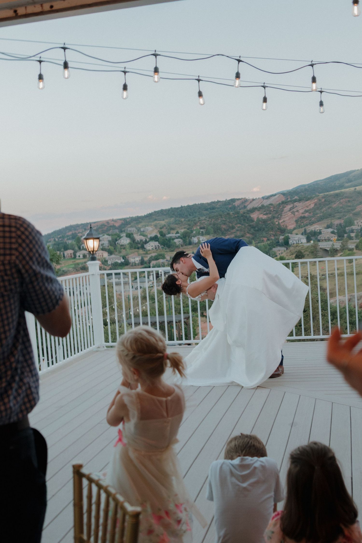 Wedding guests look out over mountain view from white deck with string lights overhead.