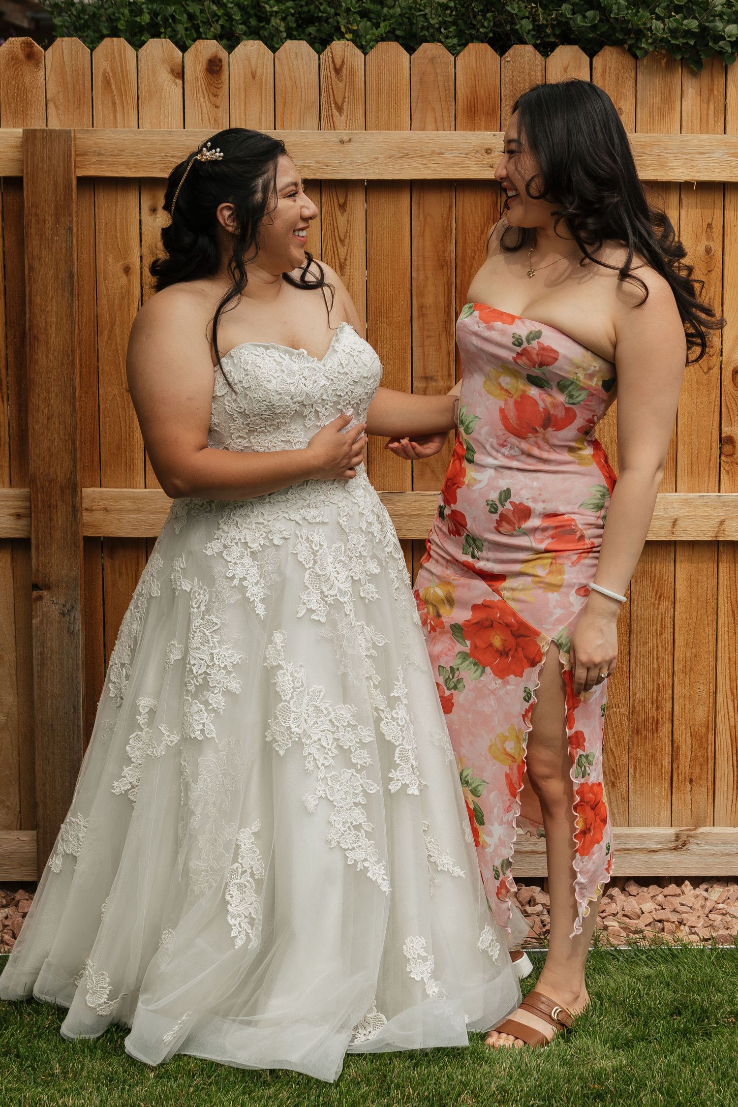 A bridal party in floral dresses and white gowns poses against a wooden fence backdrop.