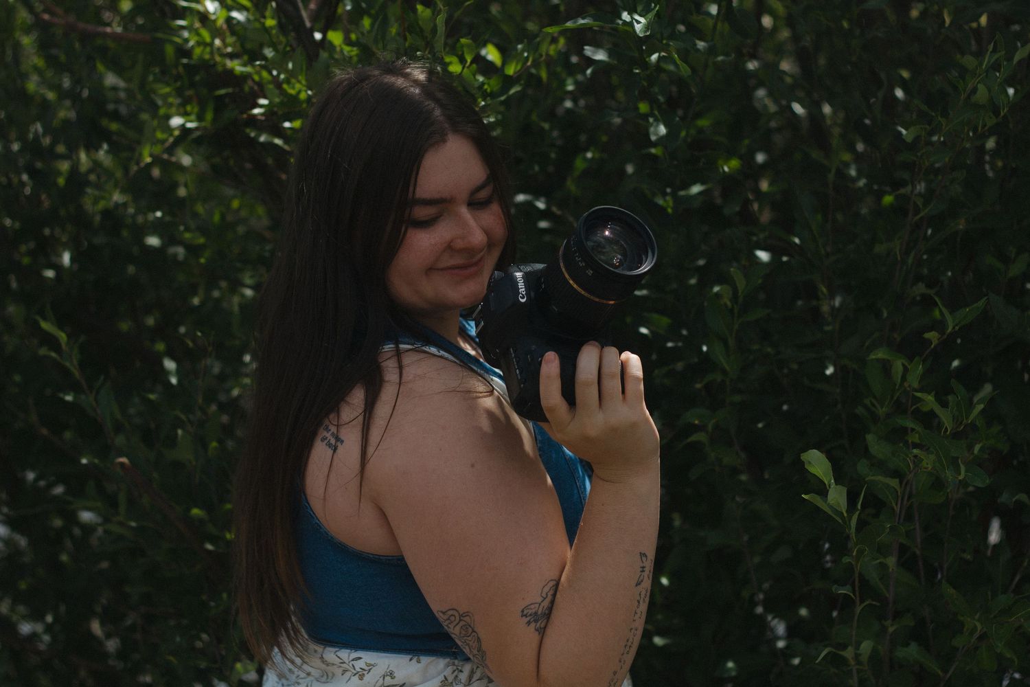 A person in a blue dress stands in front of lush green foliage while taking a selfie.