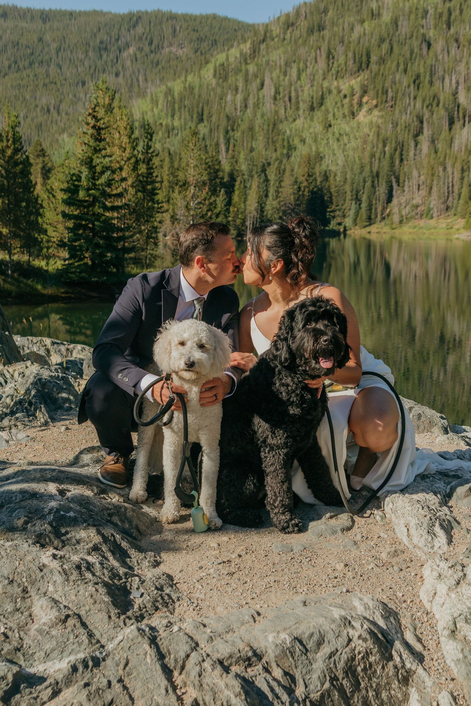 A couple shares a tender moment with their two dogs on rocky terrain by an alpine lake.