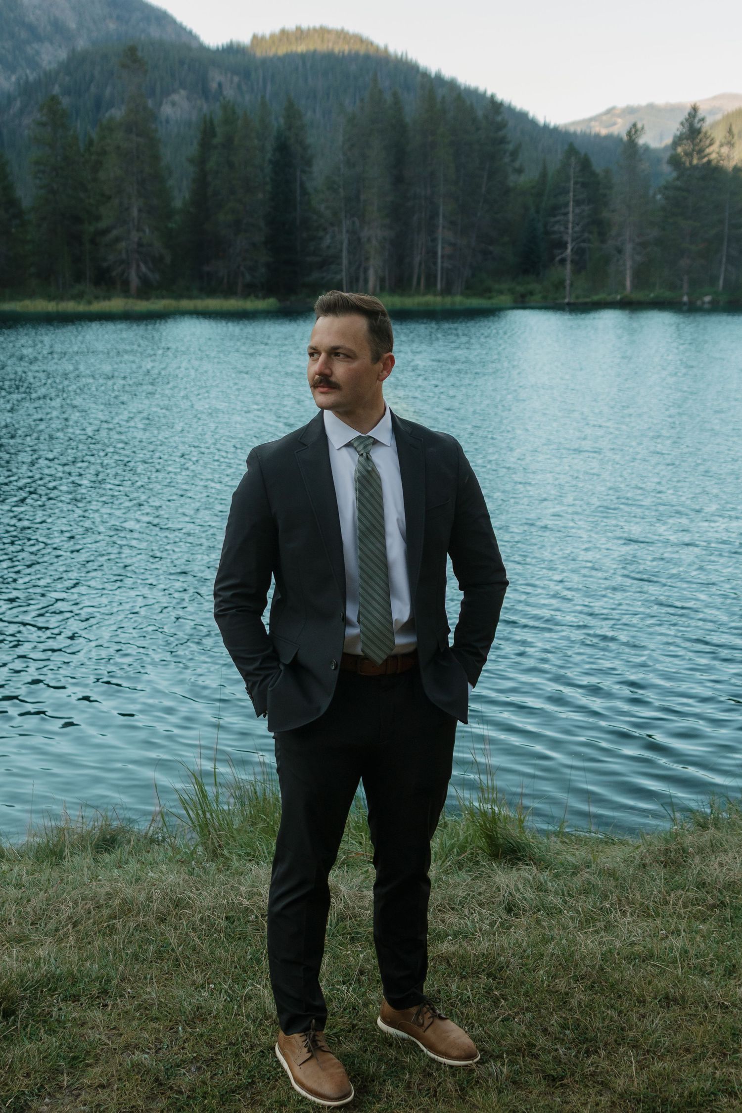 Groom in dark suit poses by serene mountain lake with evergreen forest backdrop.