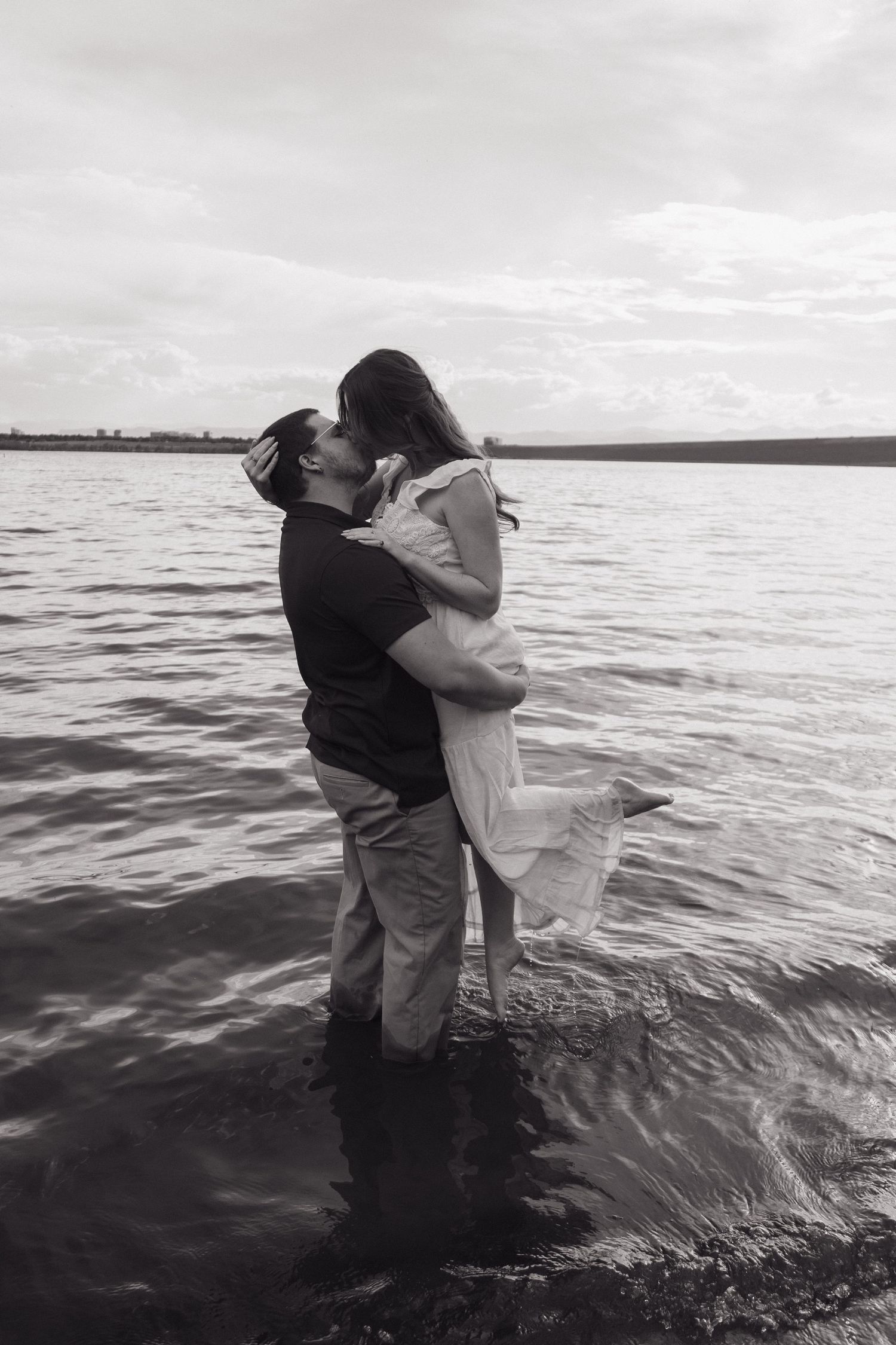 Black and white photo of a couple kissing while standing in water, the woman lifted slightly, with a serene lake backdrop.
