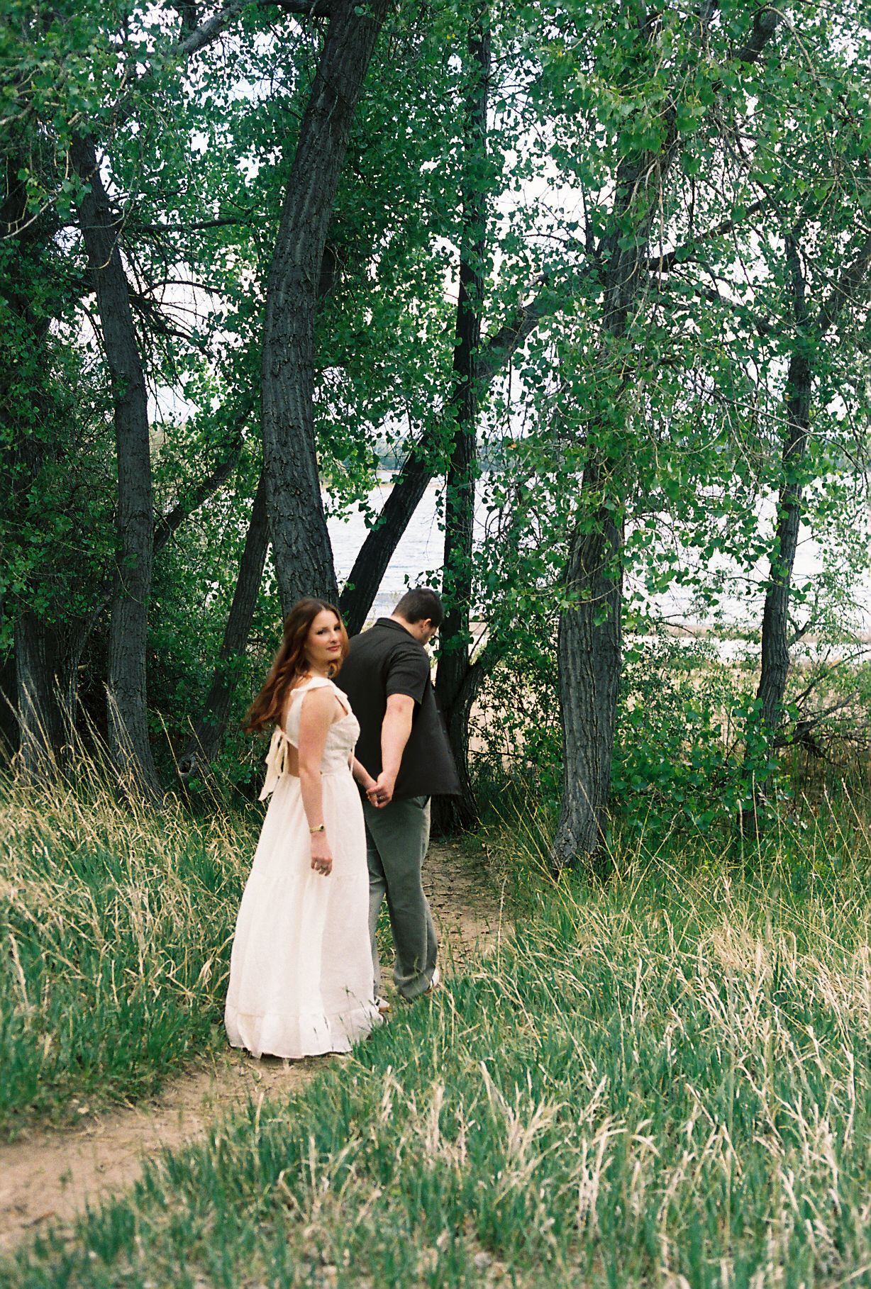 Couple walking hand in hand on grassy path through lush green trees, woman glancing back.