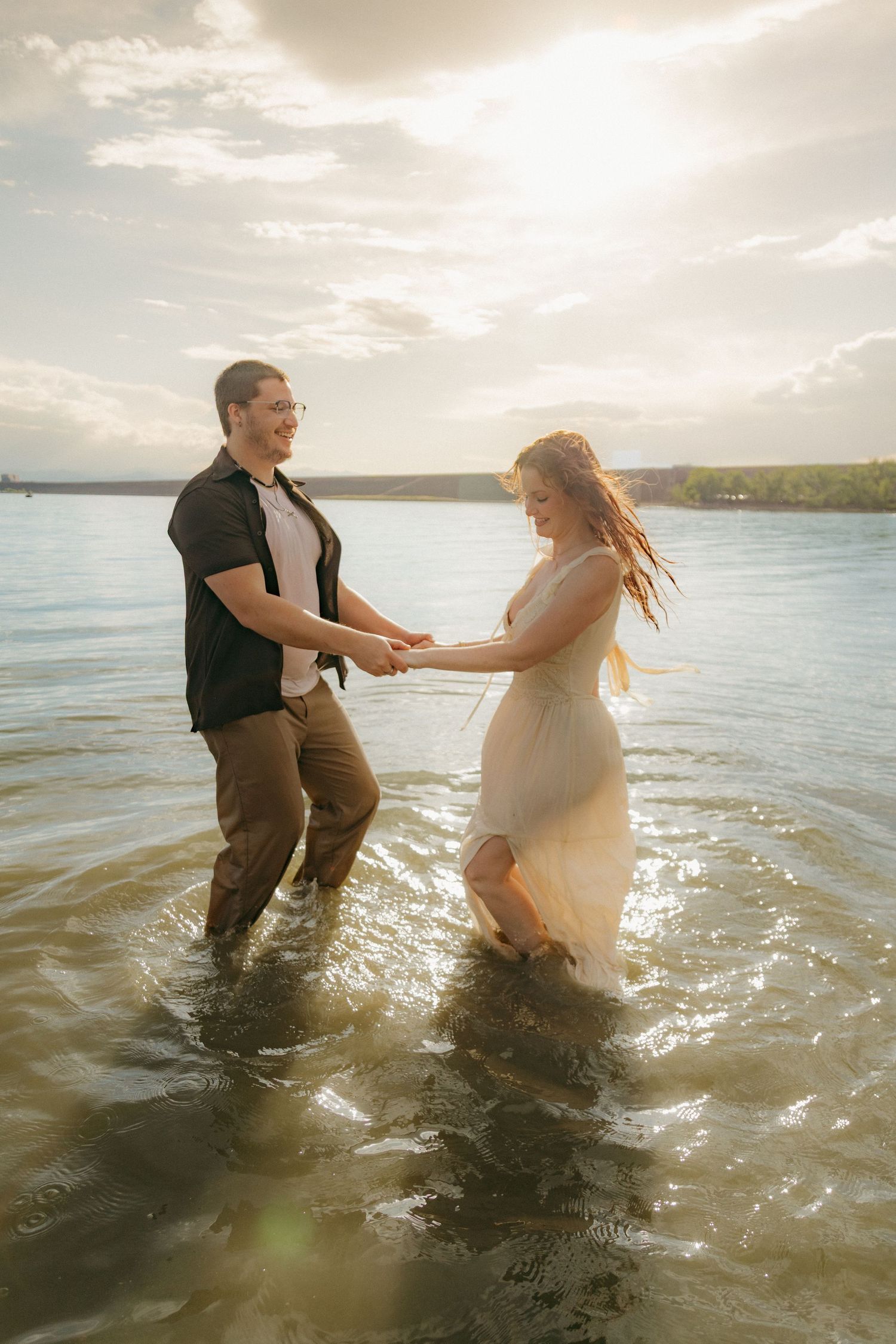Joyful couple holding hands and walking through shallow lake water at sunset.
