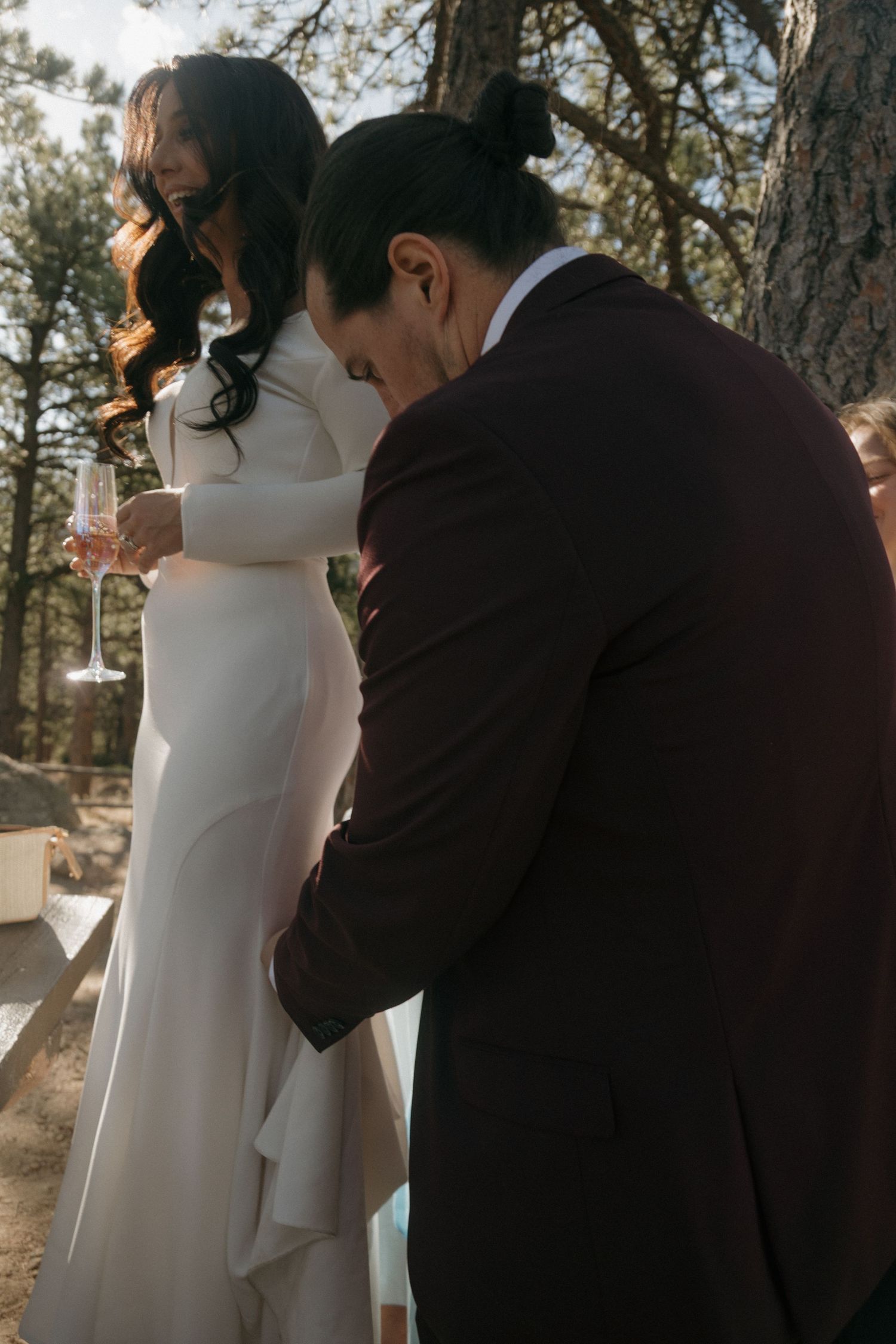 Bride in white gown holding champagne glass stands with groom in burgundy suit bowing head among pine trees.