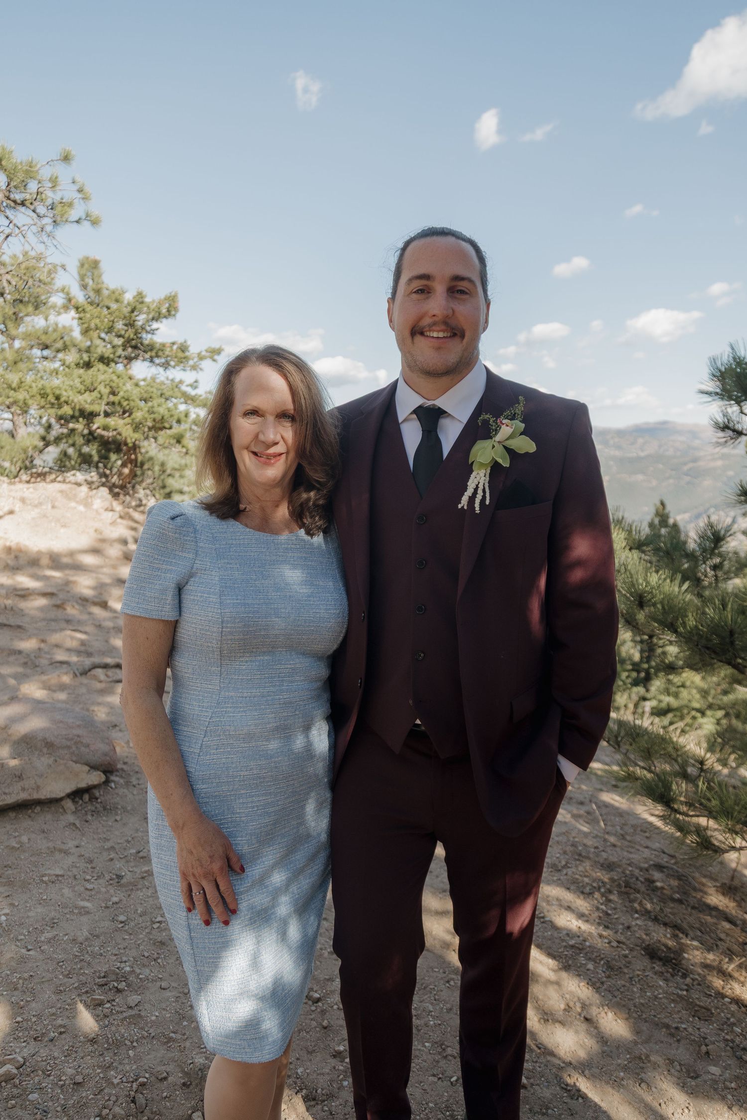 Groom in burgundy suit poses with woman in blue dress at outdoor mountain wedding.