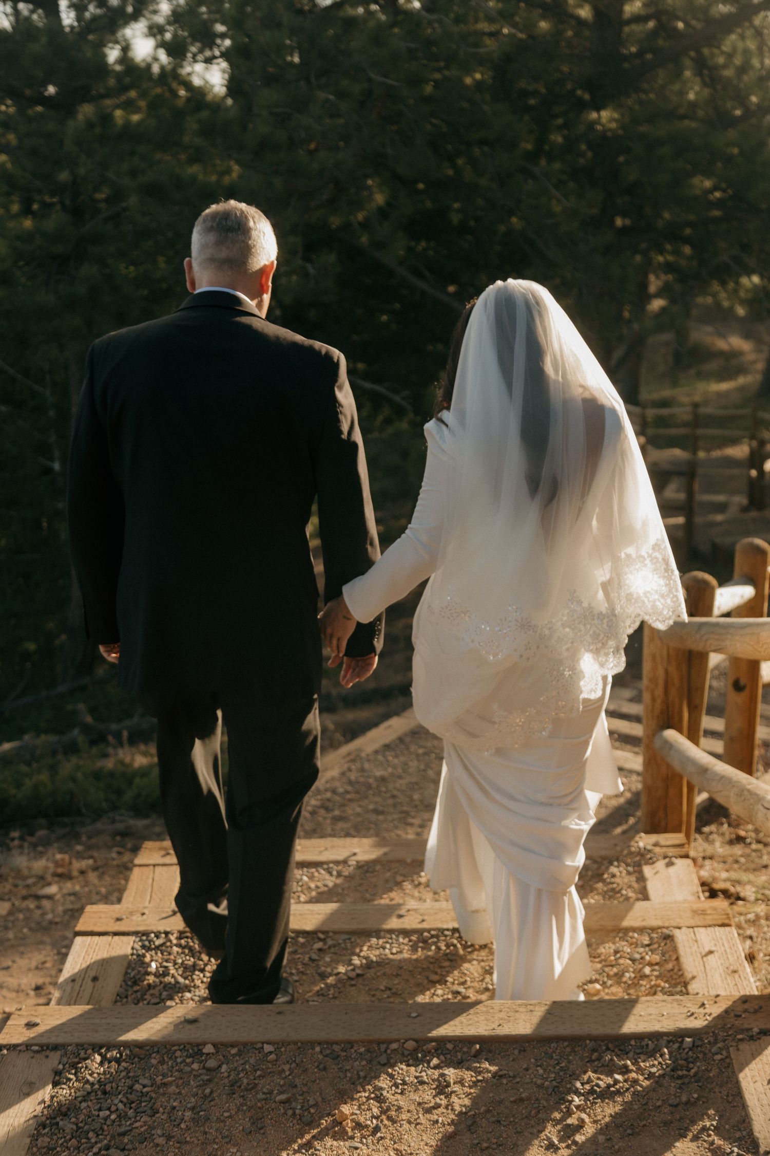 Bride and groom walk hand-in-hand up wooden steps through a sunlit forest after ceremony.