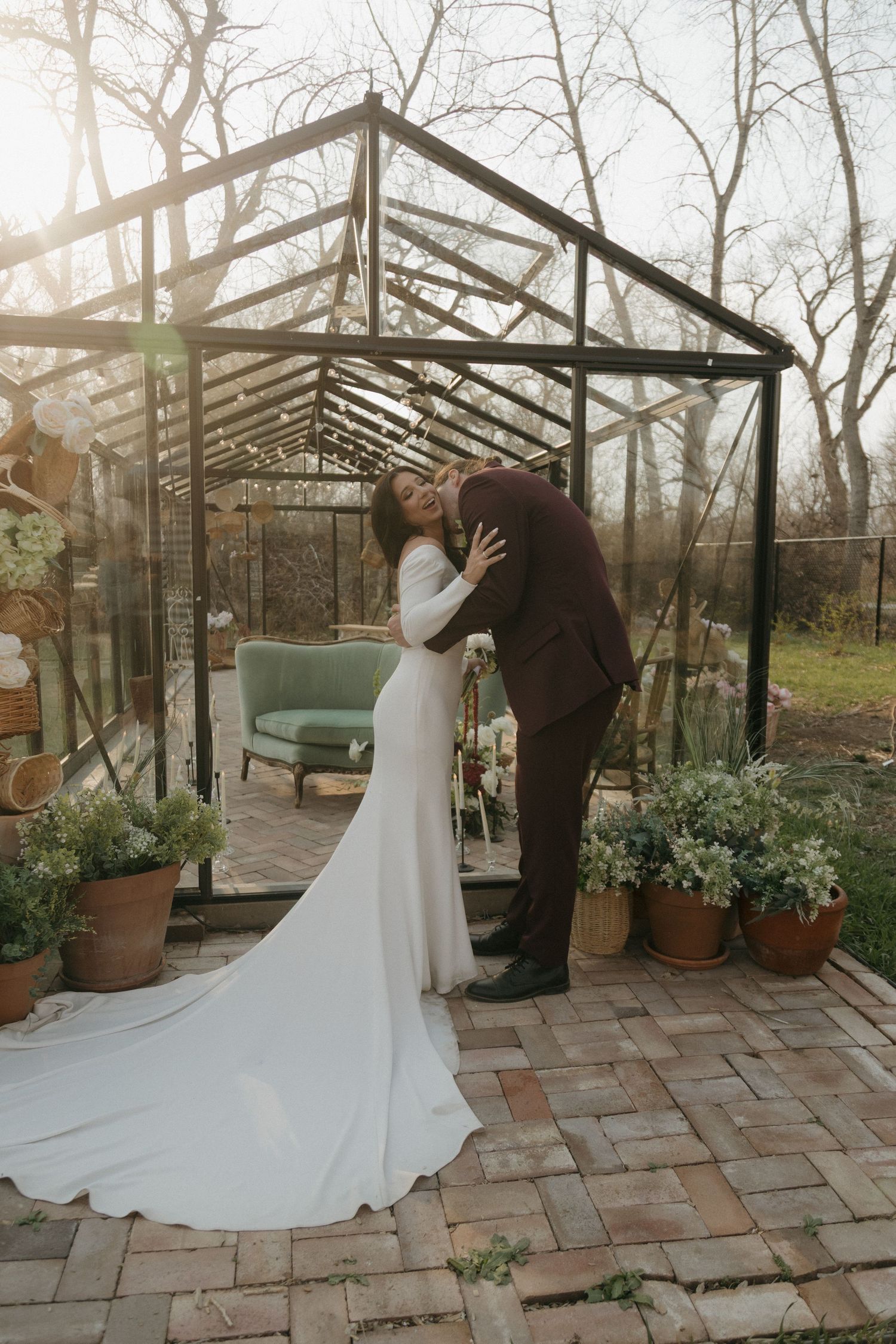 Bride and groom share a romantic embrace outside a glass greenhouse at golden hour.