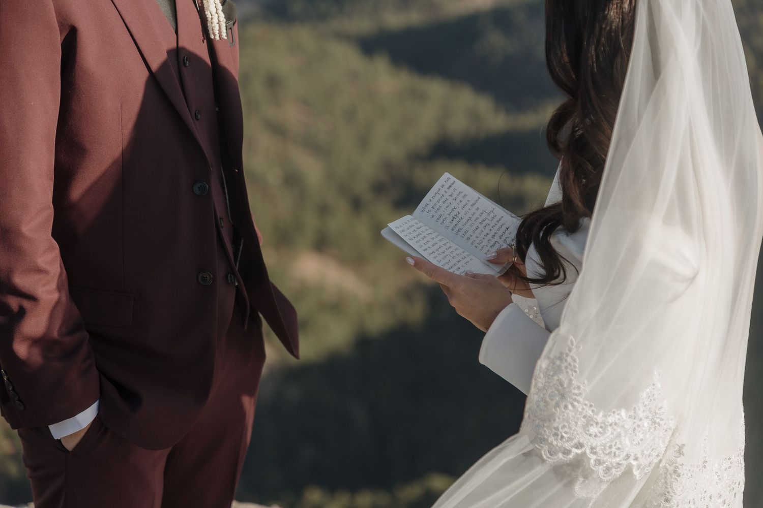 Bride in lace veil reading handwritten vows to groom in burgundy suit at mountain wedding.