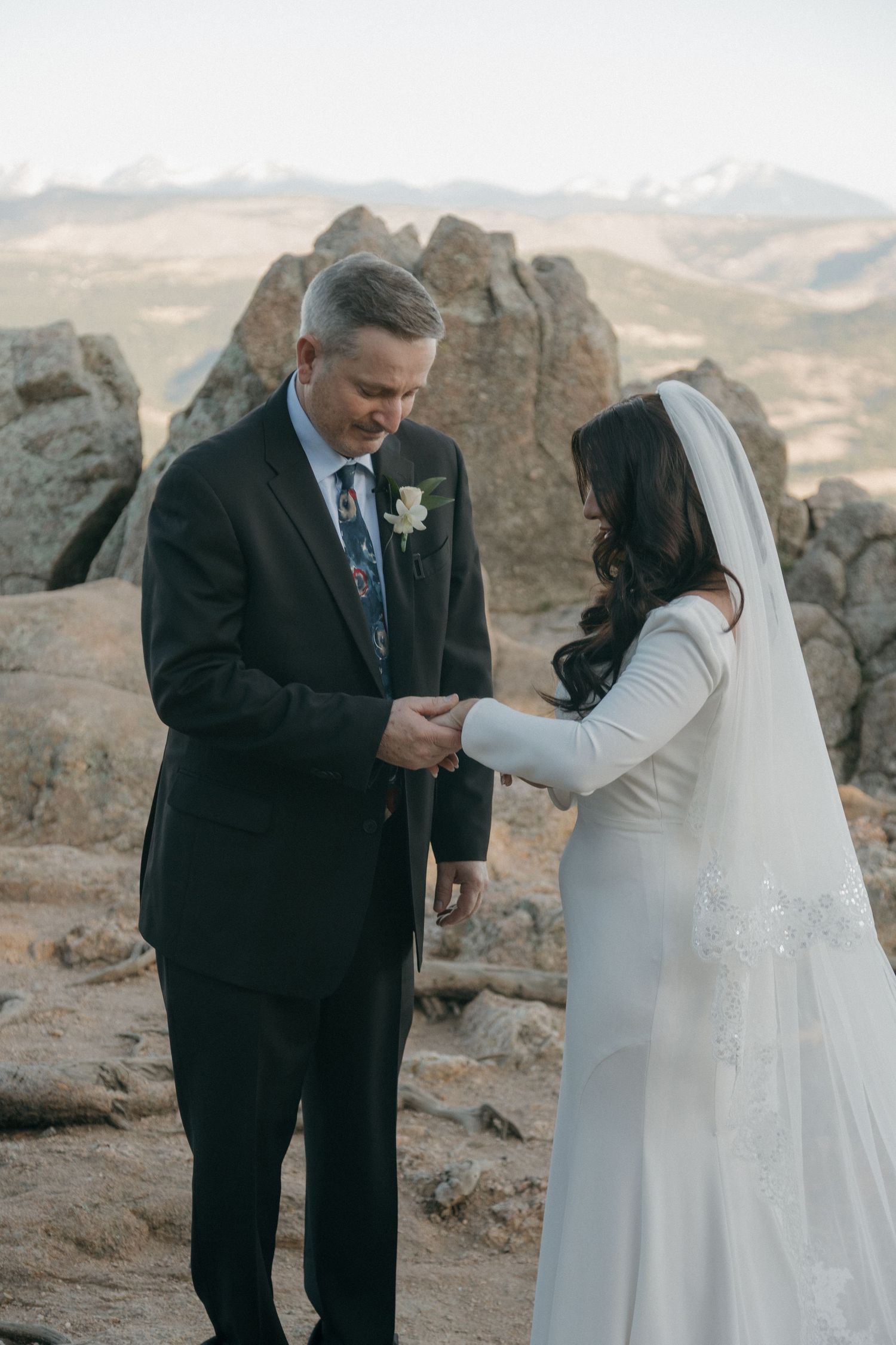 Groom in dark suit holds bride's hands during outdoor mountain elopement ceremony at Rocky Mountain National Park.