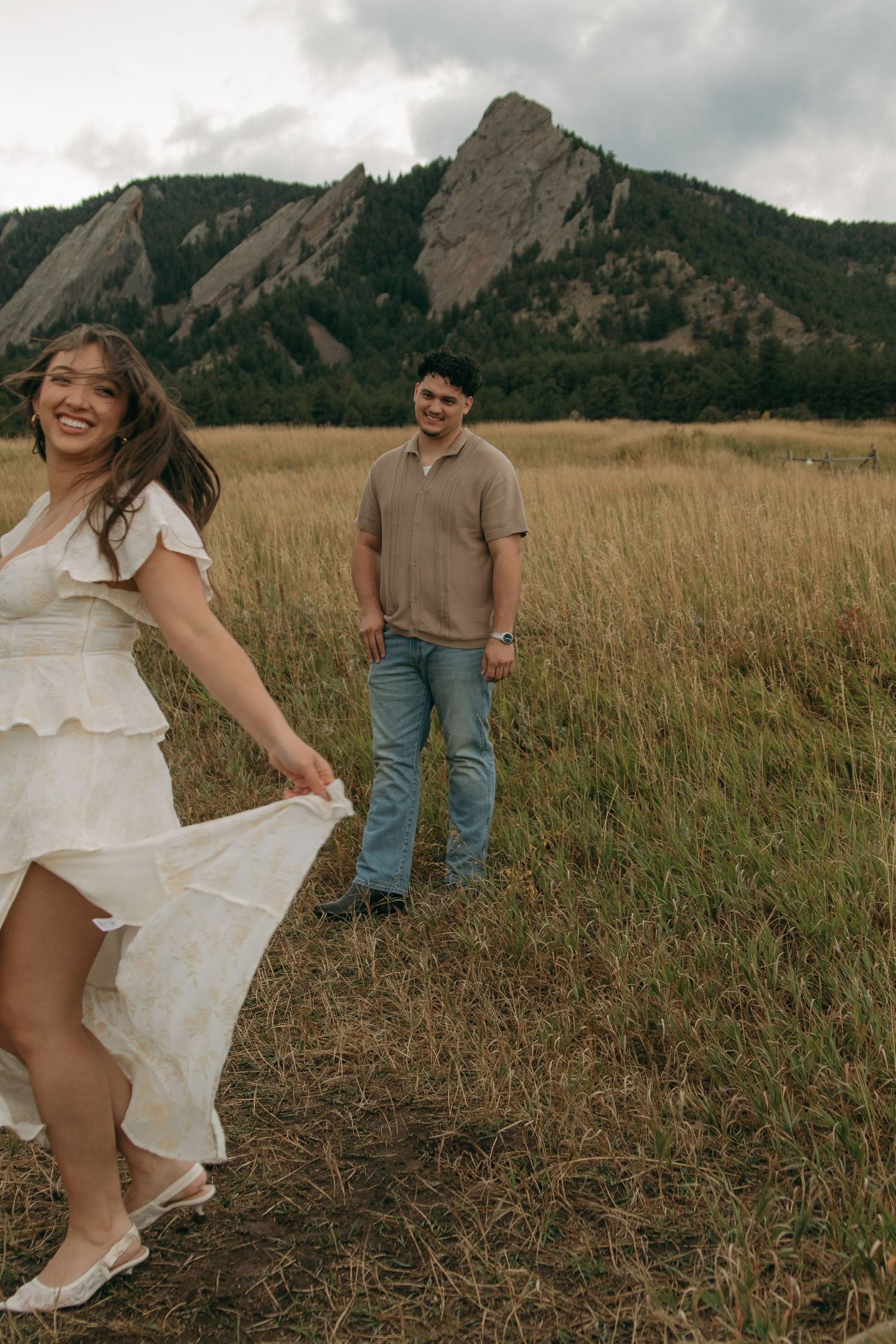 Woman in white tiered dress laughing while man watches her in a field near the Flatirons.
